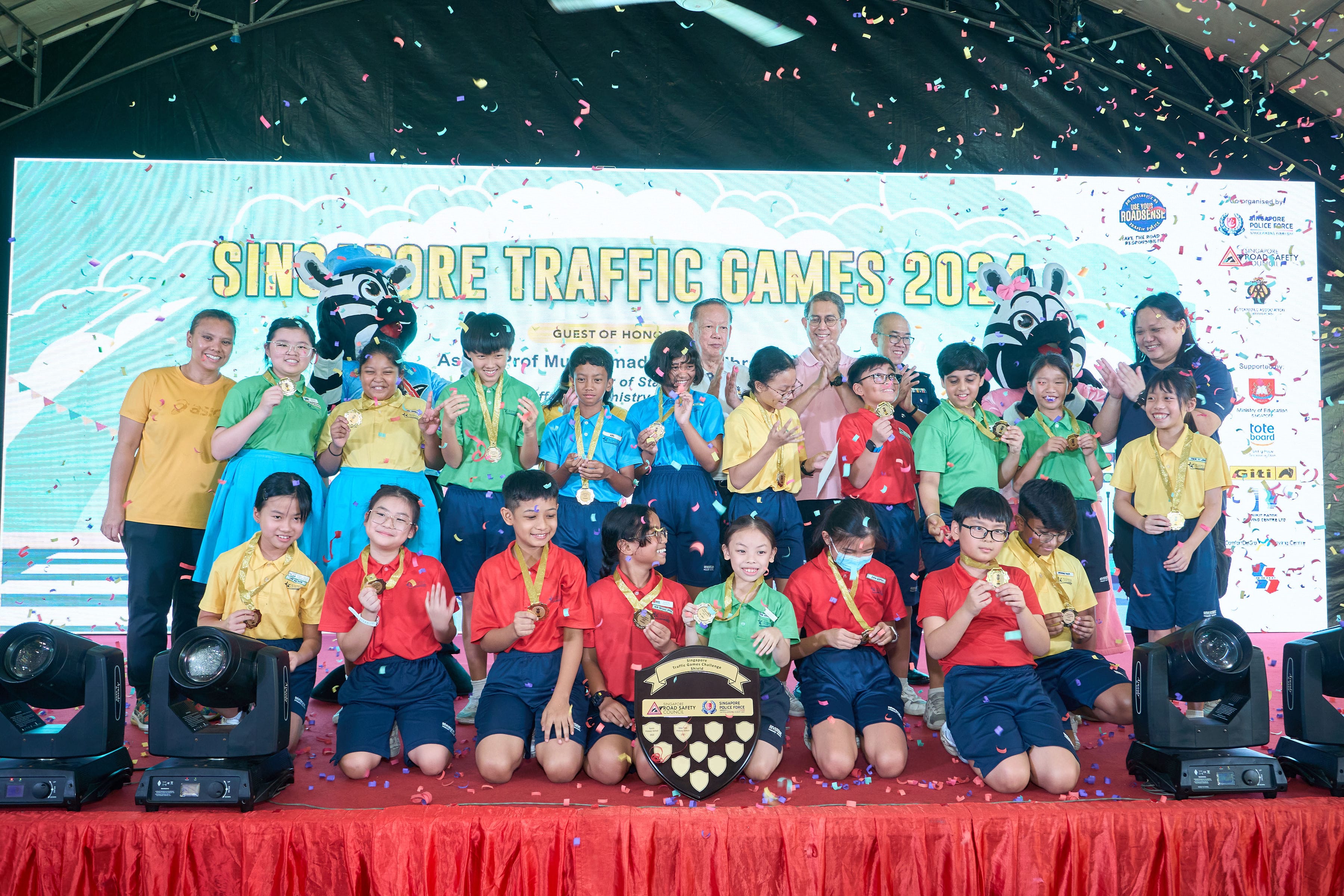 A group of children in colorful uniforms and medals stand on a stage with confetti, a large trophy, and event signage behind them.