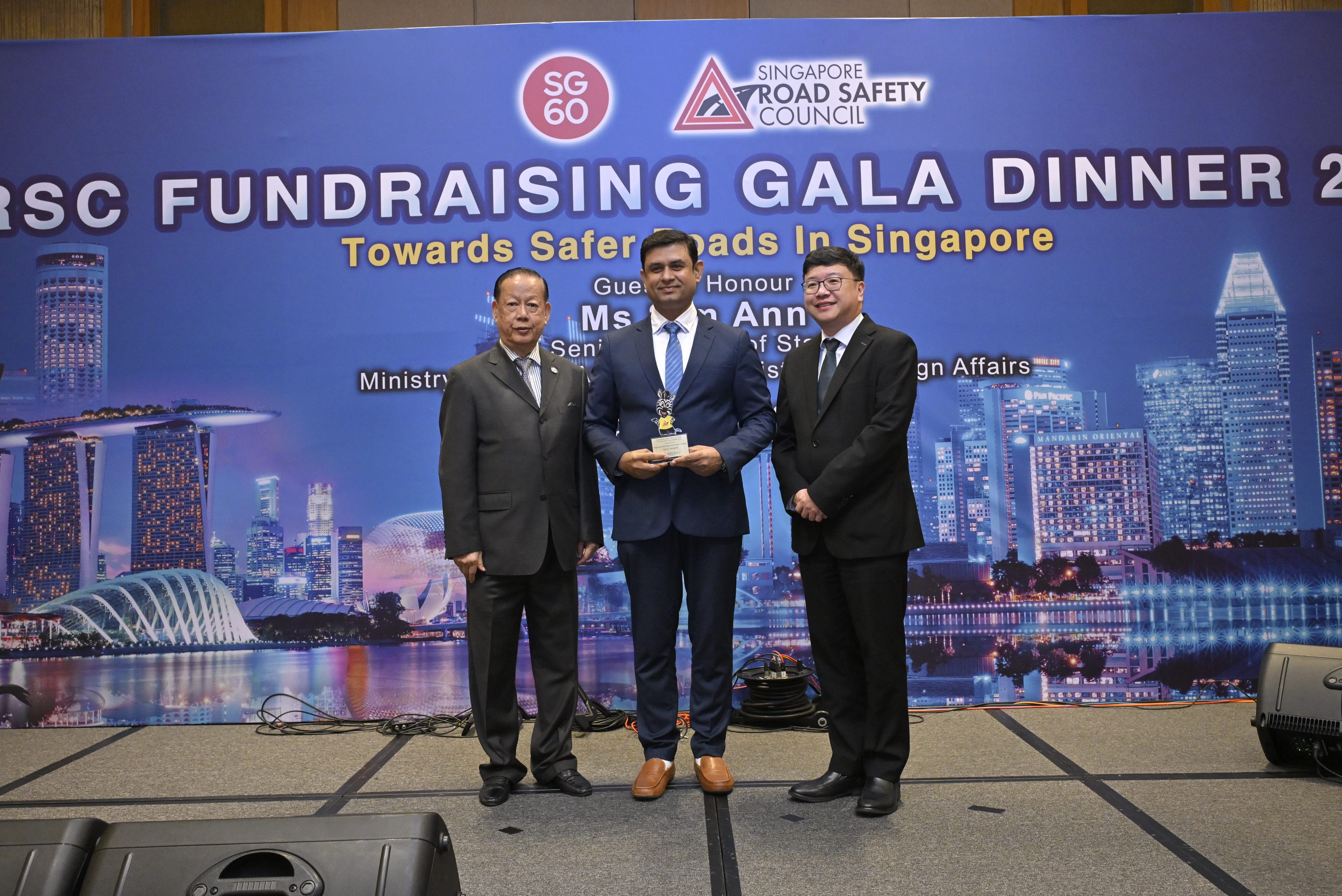 Three men in suits stand on stage at Singapore Road Safety Council fundraising gala, one holding an award.