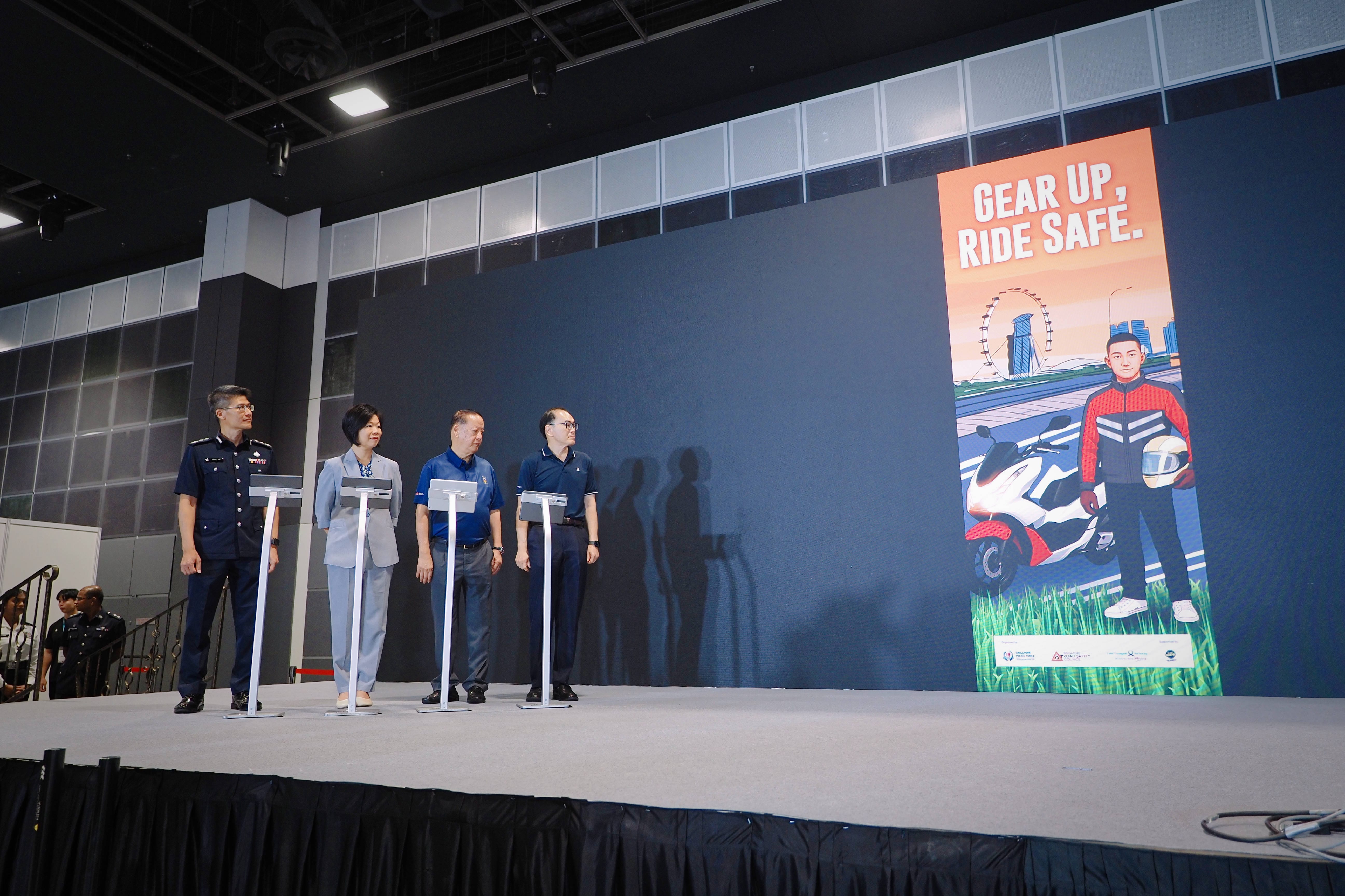 Four people stand on stage near podiums beside a safety poster with the text "Gear Up, Ride Safe" and an image of a motorcyclist.