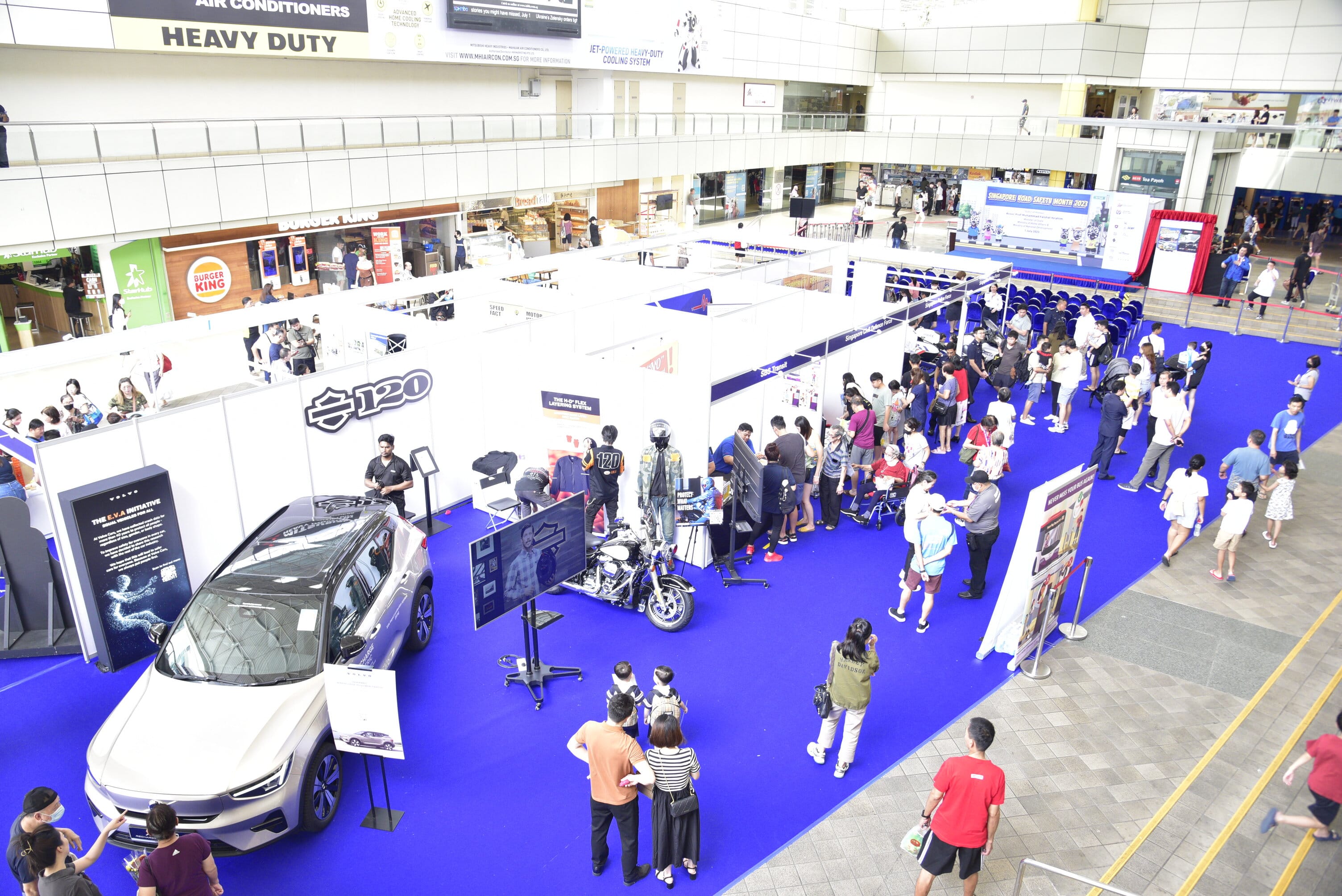 Exhibition hall with car and motorcycle displays, numerous attendees, booths, and seating area against a blue carpet backdrop.