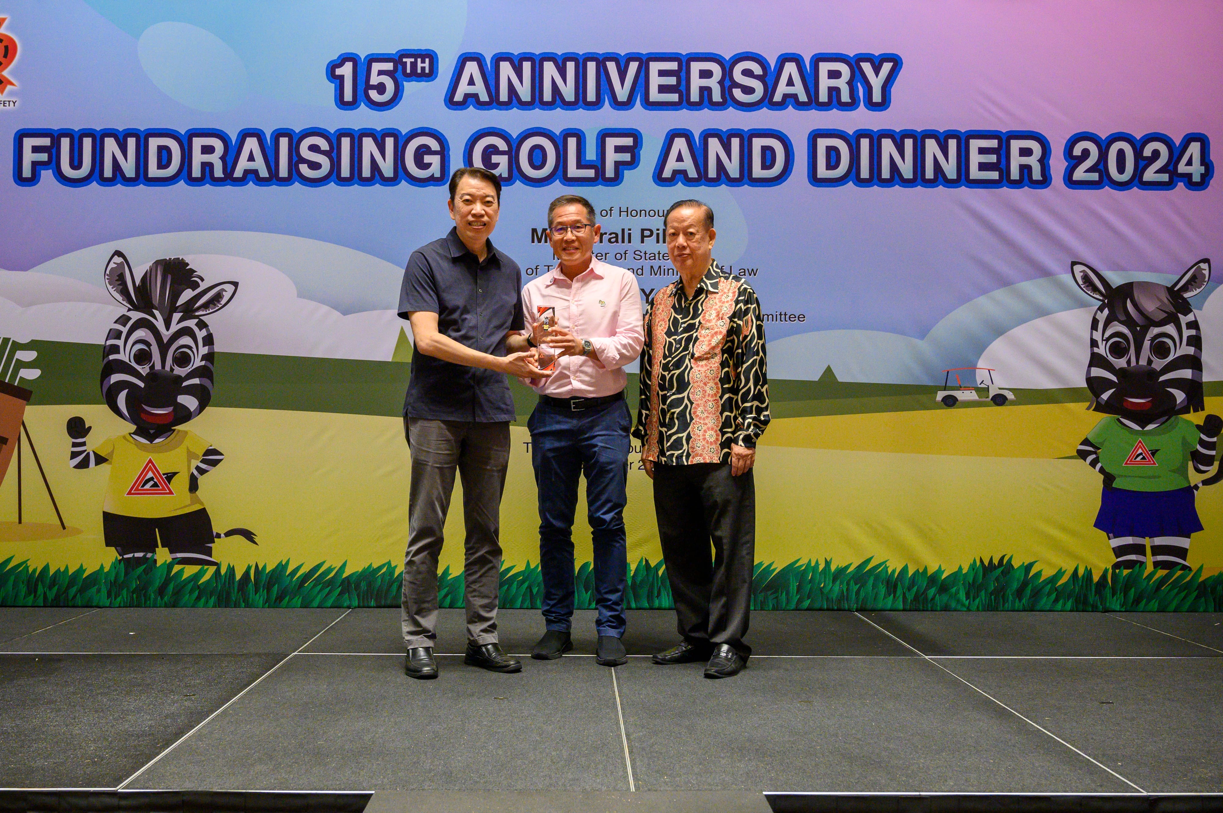Three people stand on stage at a golf and dinner event with cartoon zebras decorating the backdrop.