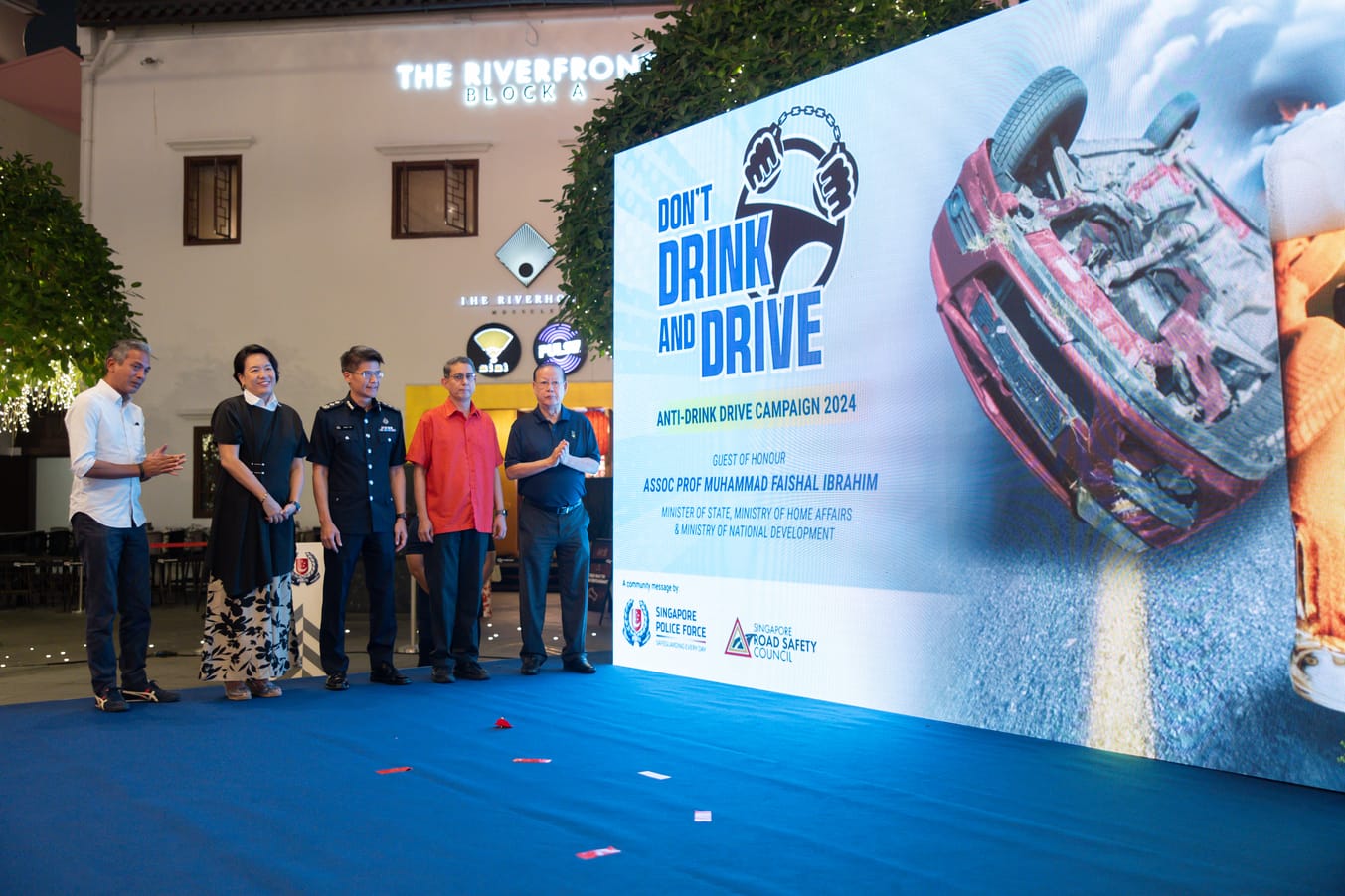 Five people standing on a blue floor next to a large sign for the "Don't Drink and Drive" campaign showing a car accident.