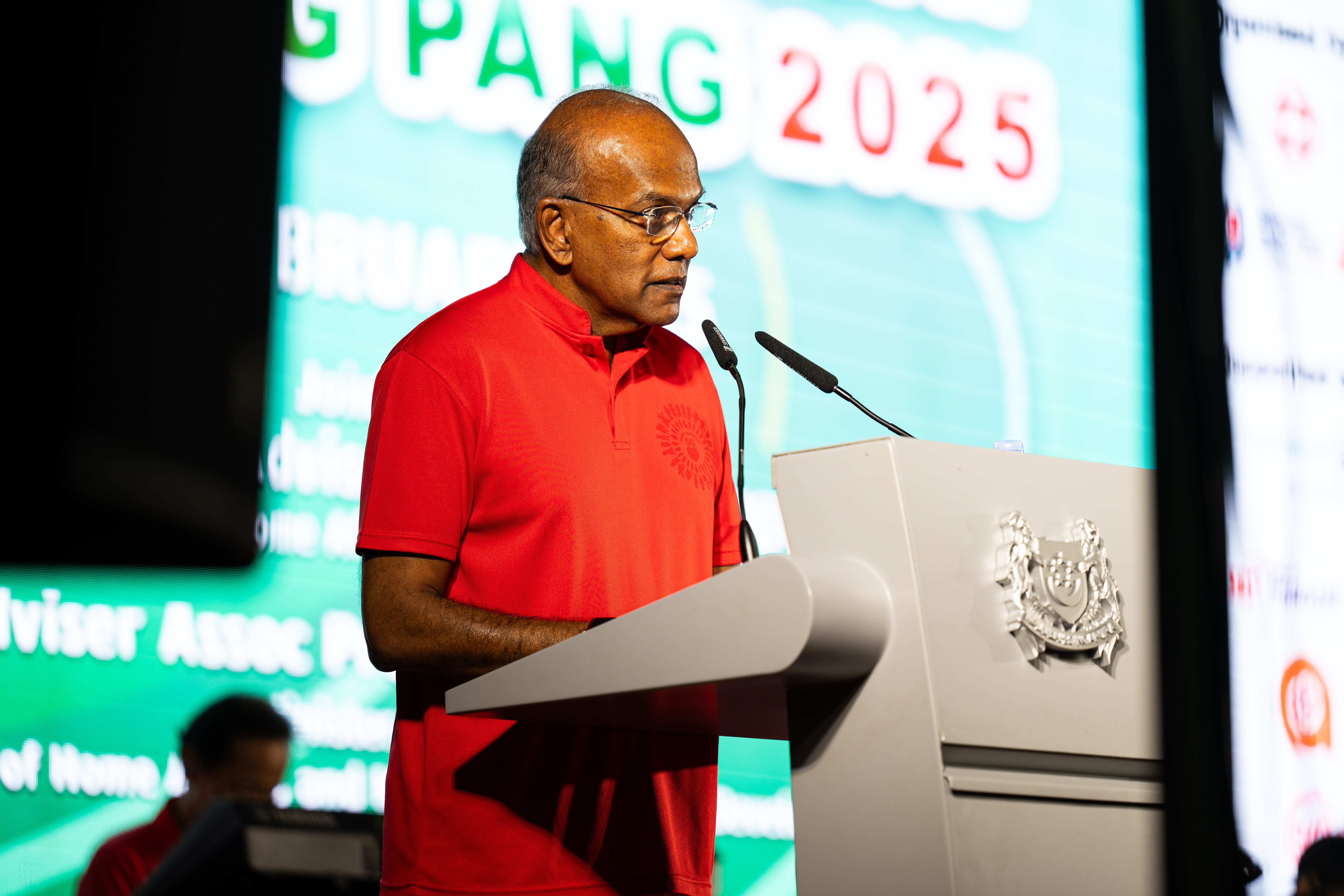 A person in a red shirt speaks at a podium with a crest, words "PANG 2025" visible on the screen behind them.