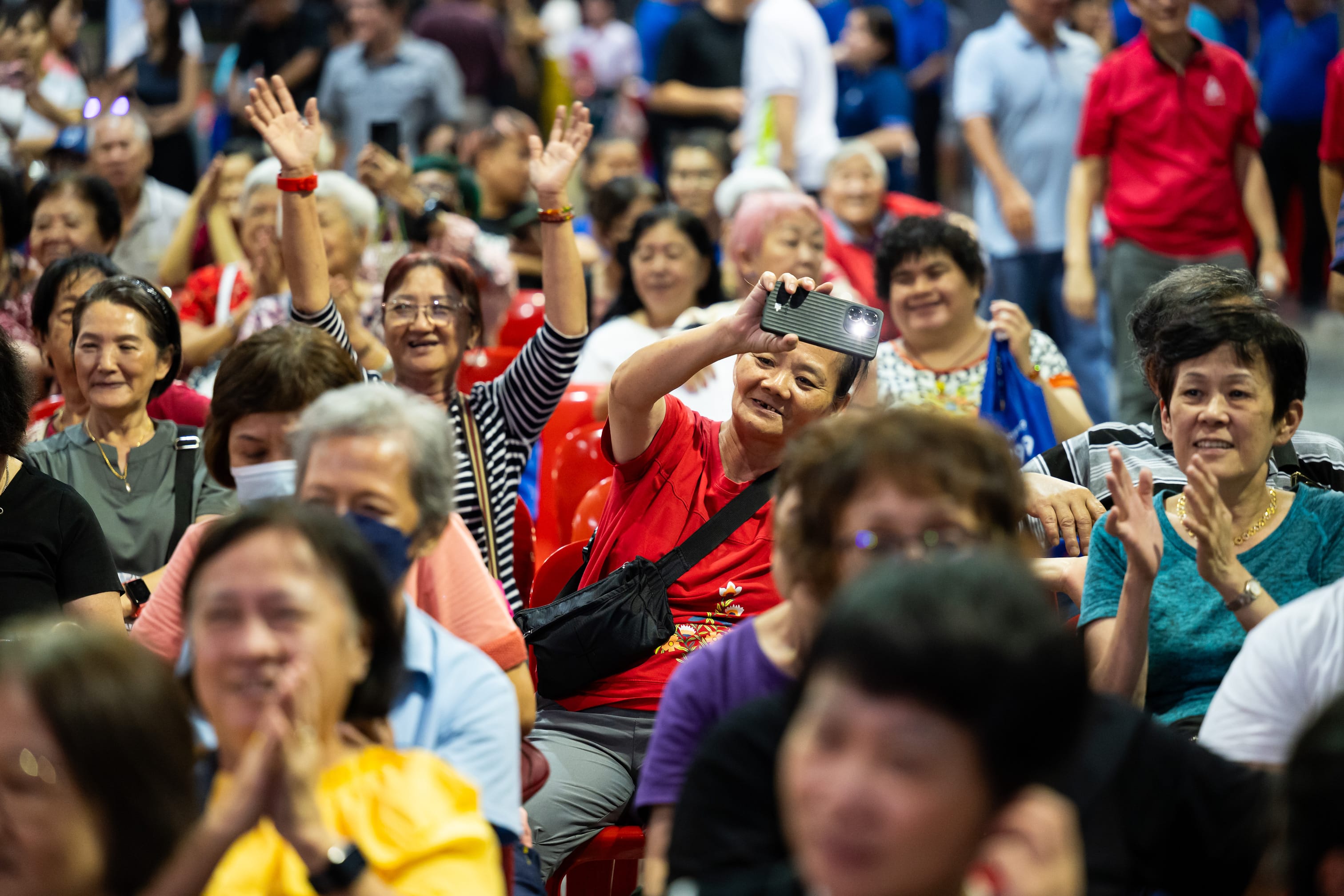 People seated in red chairs in a crowded indoor setting, some raising their arms and another taking a photo.