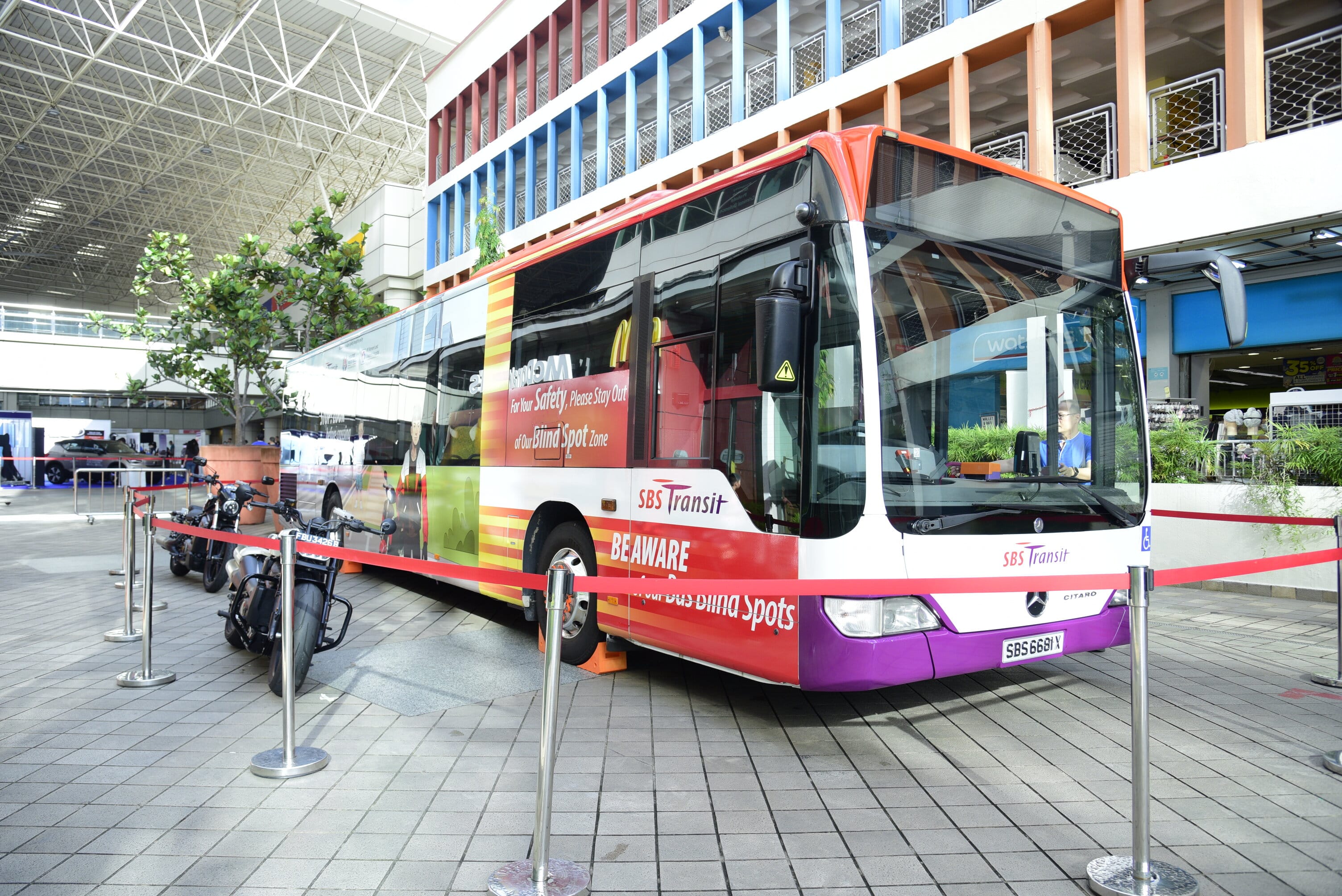 A large city bus with "SBS Transit" branding is parked indoors near bikes and surrounded by red tape barriers.
