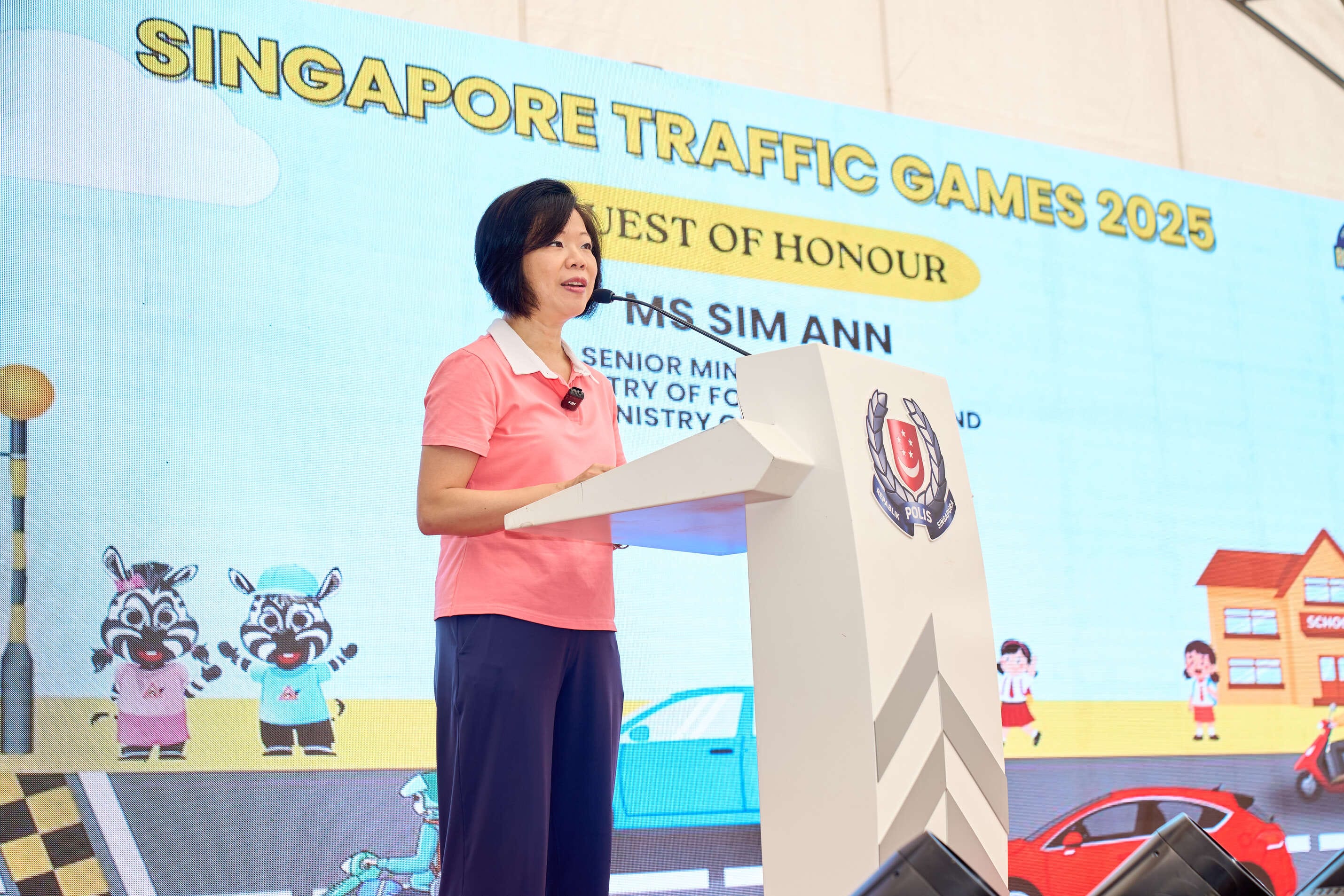A person in a pink polo shirt speaks at a podium with the Singapore Traffic Games 2025 banner behind them.