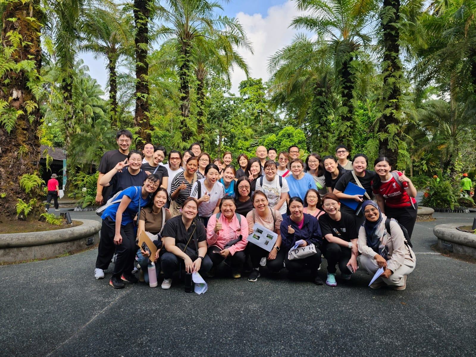 A group of people posing together outdoors surrounded by tall palm trees and lush greenery on a paved walkway.