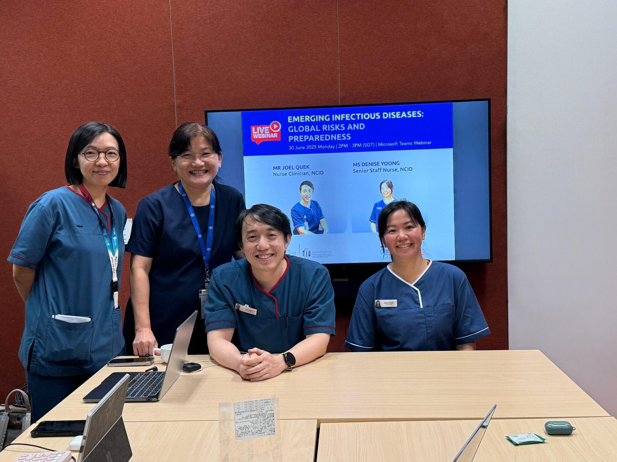 Four people in blue medical uniforms are in a room with a large screen displaying a webinar on infectious diseases.