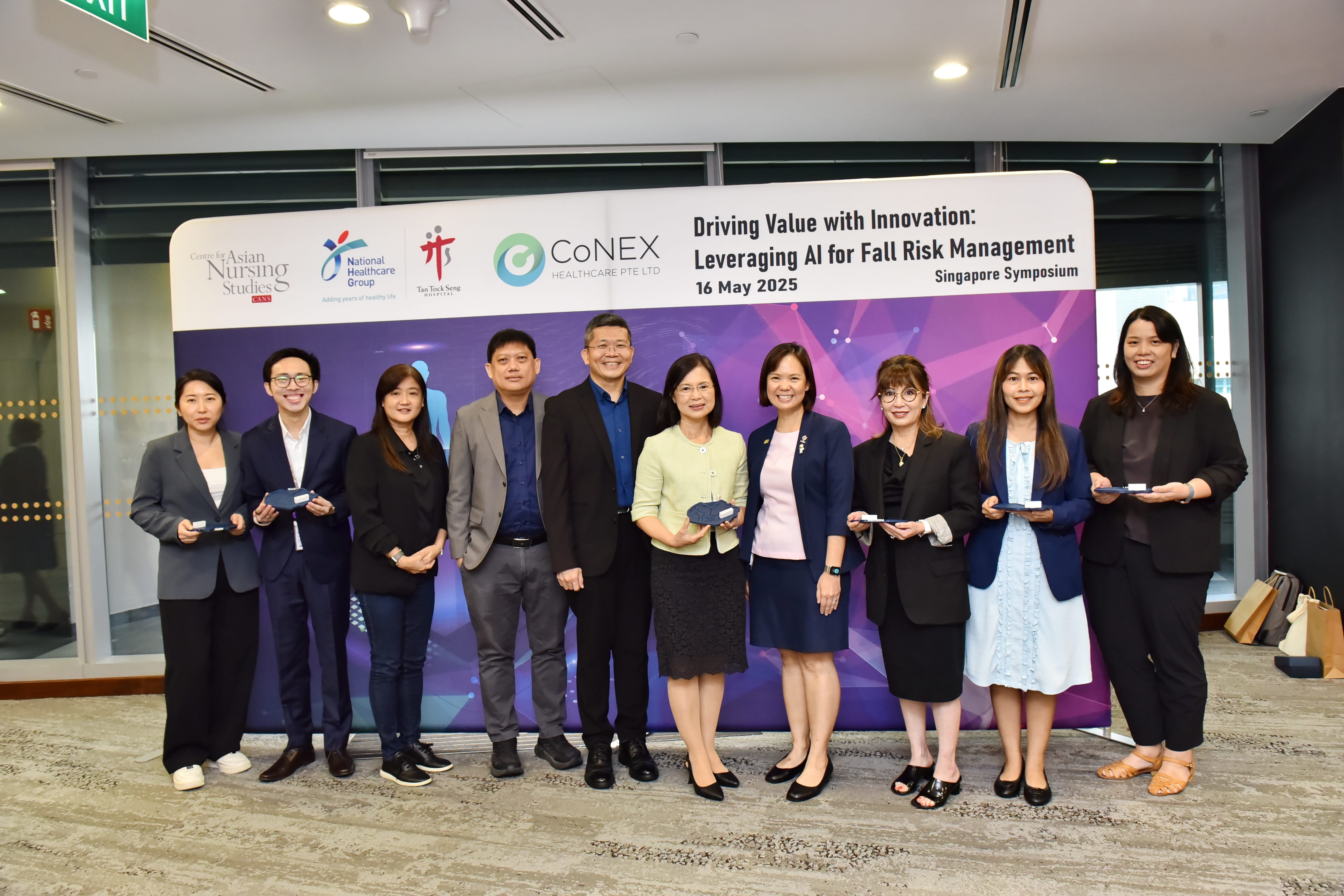 A group of people standing in front of a billboard about a symposium on leveraging AI for fall risk management dated May 16, 2025.