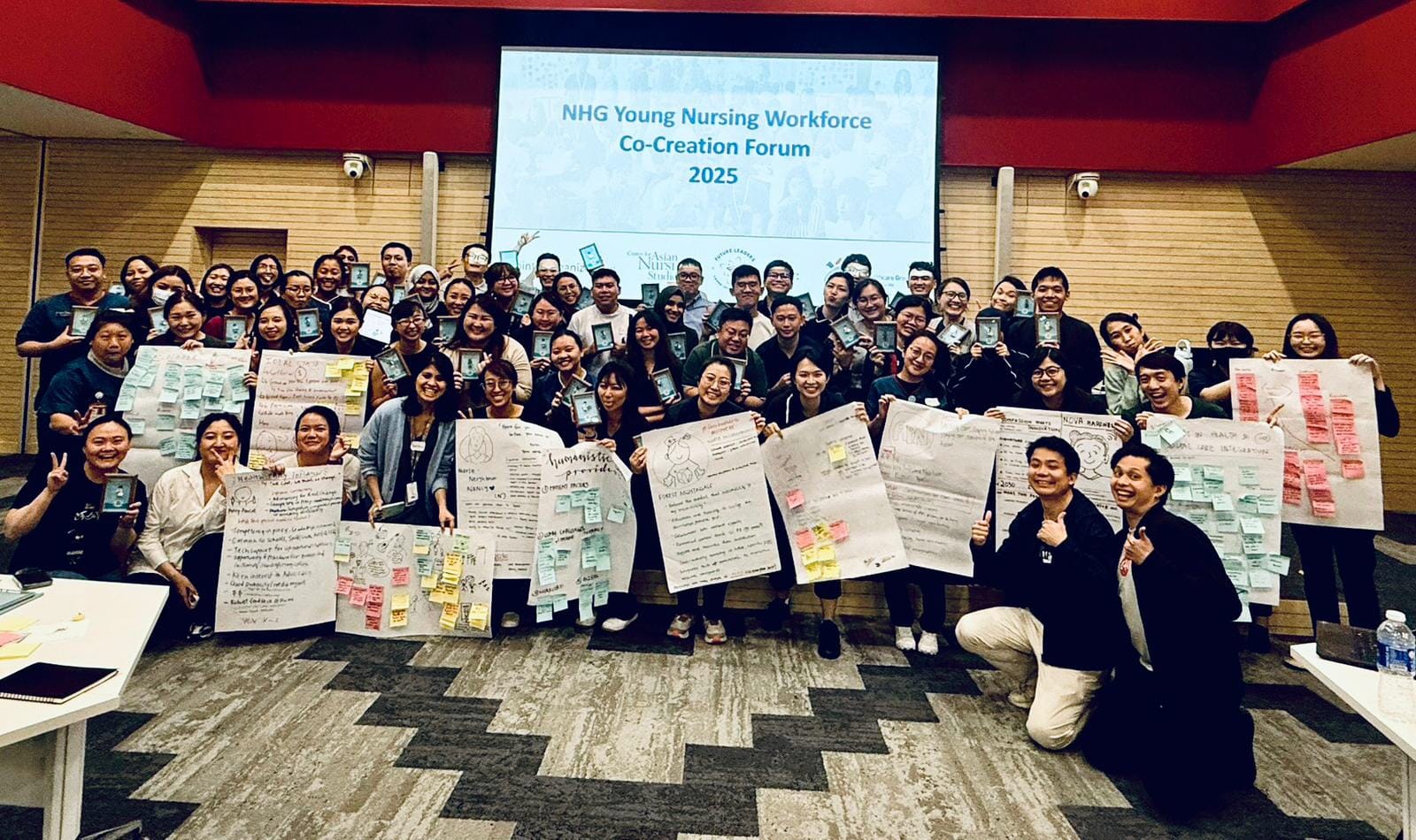 A large group of people holding posters and notes stands in front of a screen that reads, "NHG Young Nursing Workforce Co-Creation Forum 2025".