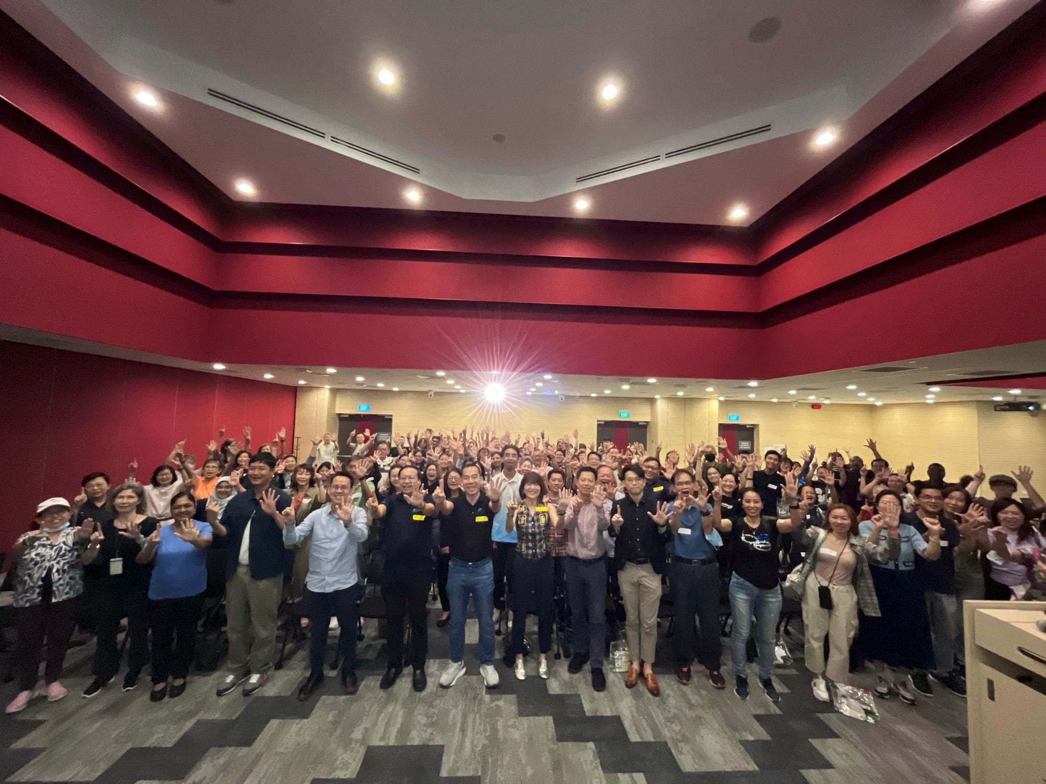 A large group of people in a conference room, many raising their hands, with red walls and bright ceiling lights.