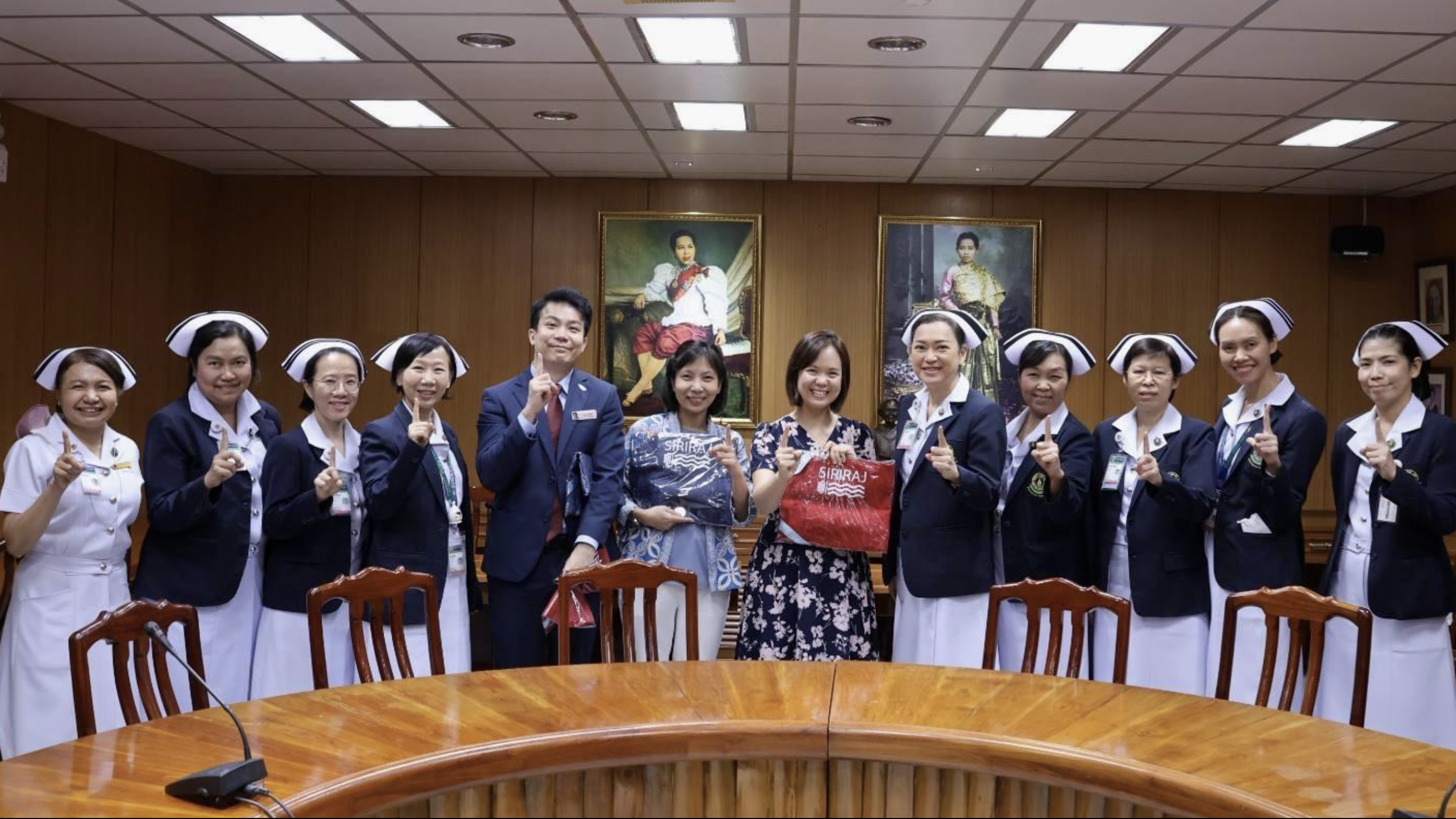 A group of nurses in uniform stand together in a room with wooden walls, holding up index fingers in front of two portraits.
