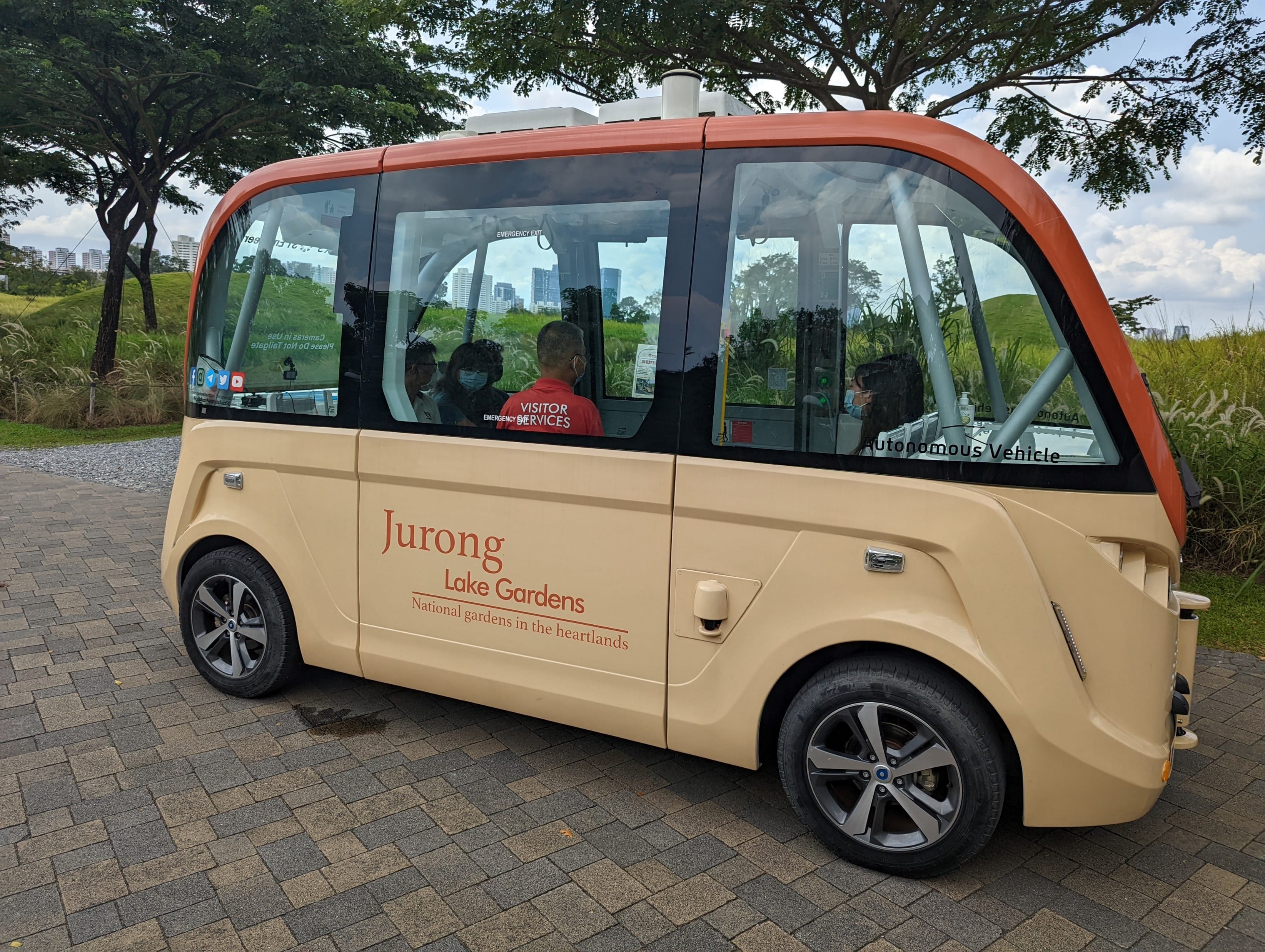 A beige autonomous vehicle labeled "Jurong Lake Gardens" with passengers inside, parked near greenery.