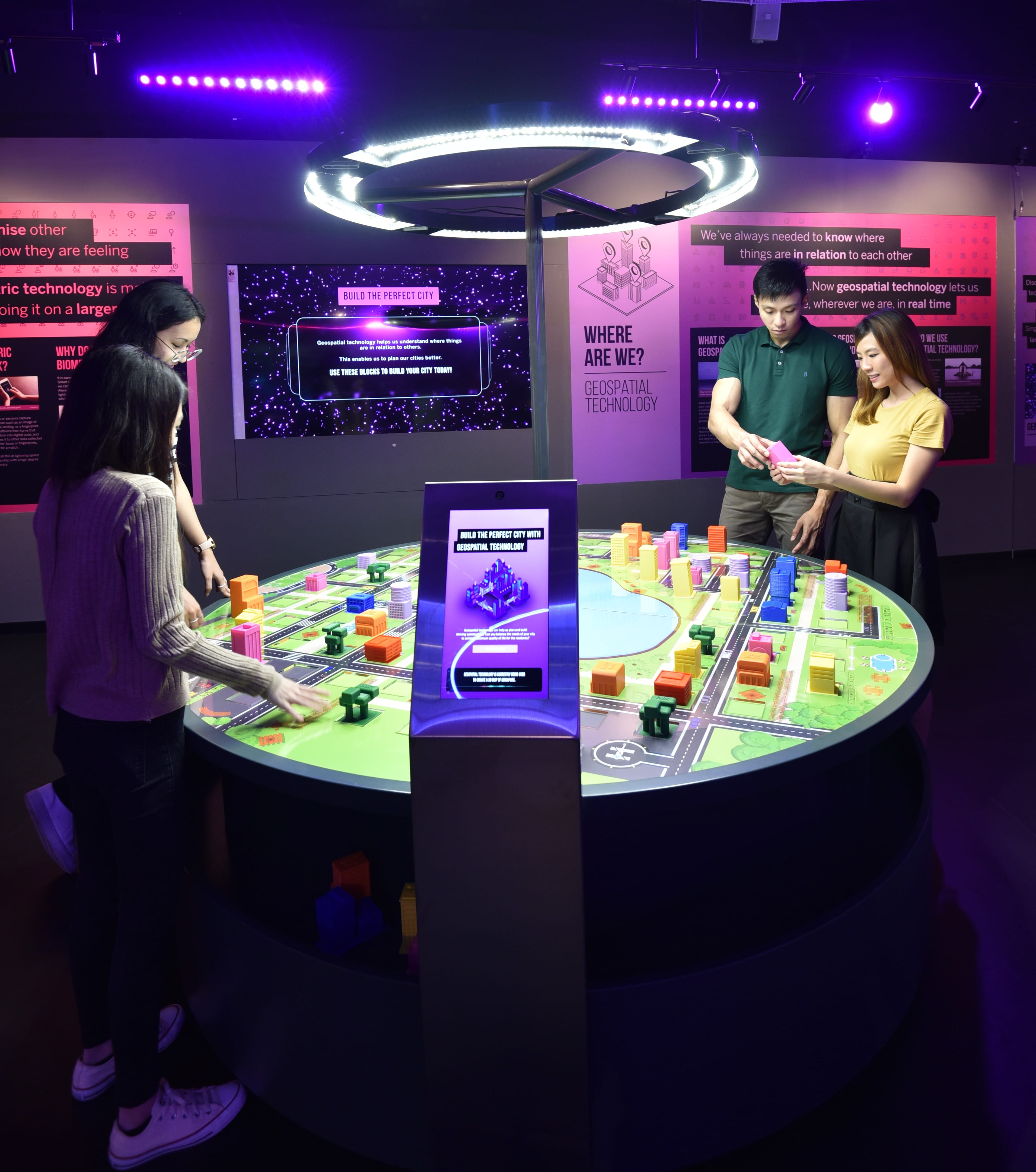 Four individuals interact with an illuminated table model of a city under purple lighting in an exhibition space.