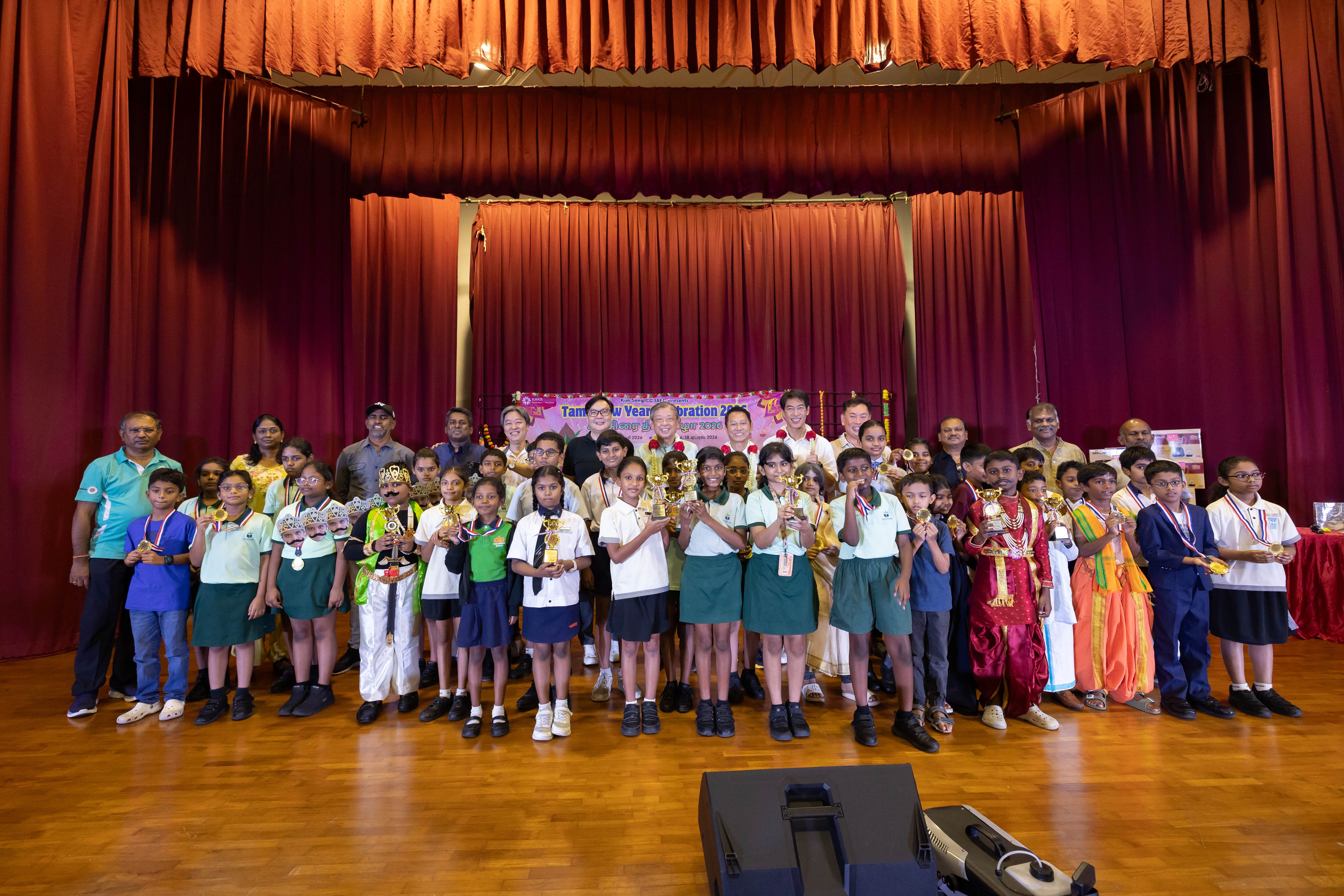 Group of children and adults pose with trophies on stage at a Tamil New Year celebration.