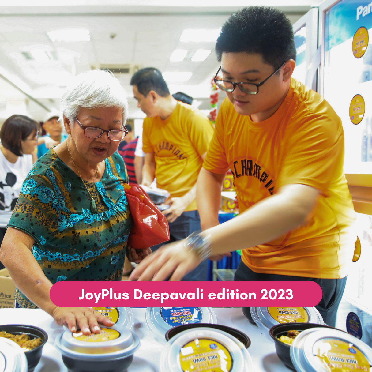 Volunteer assists senior selecting food items at JoyPlus Deepavali event.