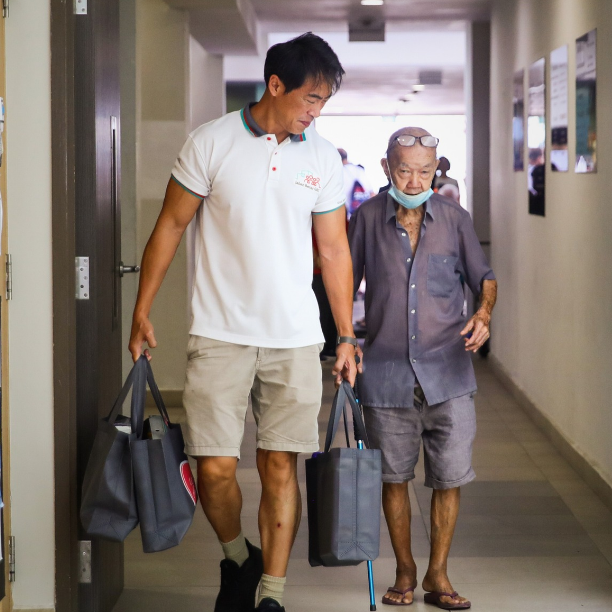 Volunteer accompanies senior carrying care packs along a residential corridor.
