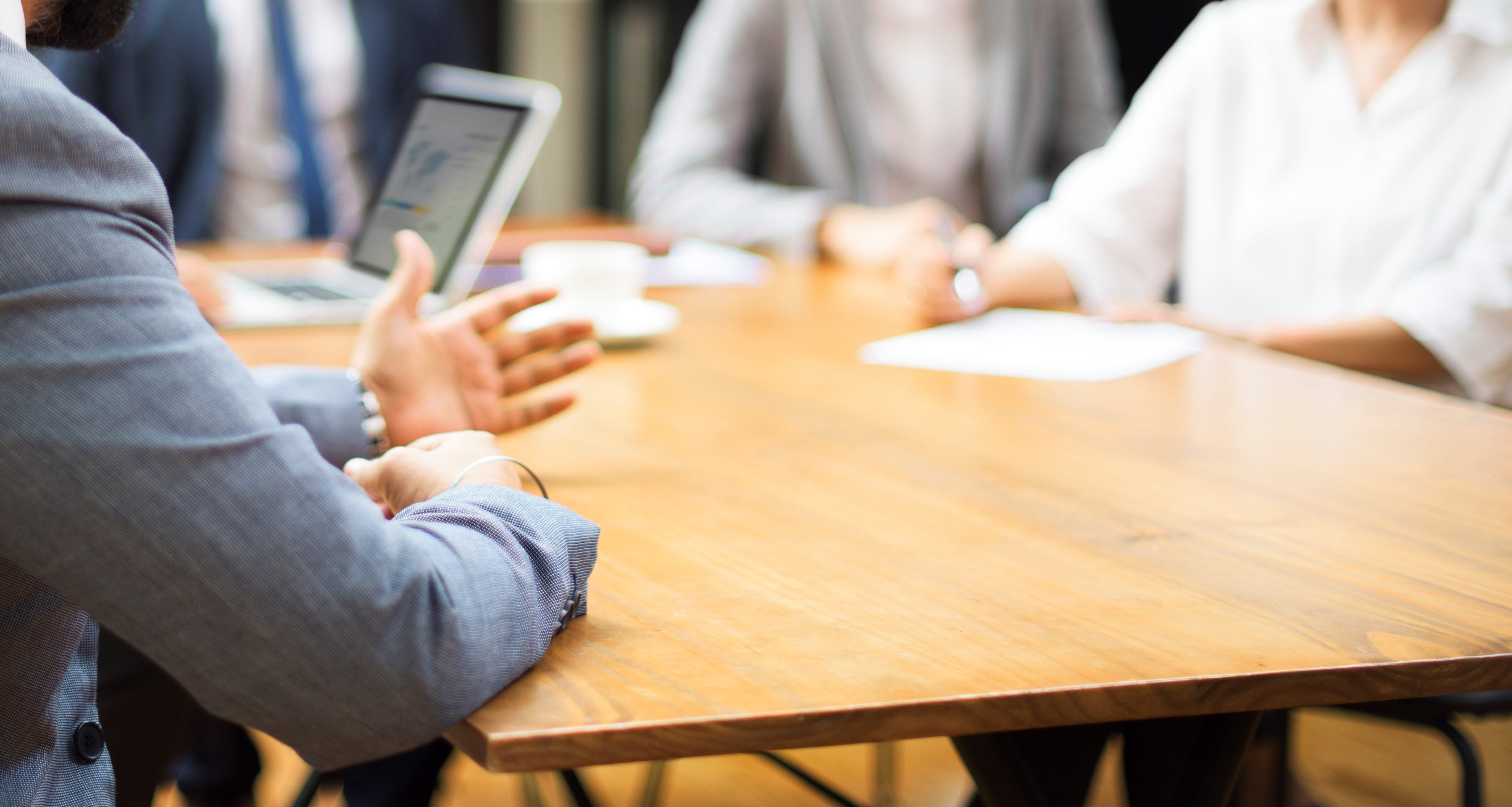 People sit at a table with laptops, papers, and a coffee cup, engaging in a discussion.