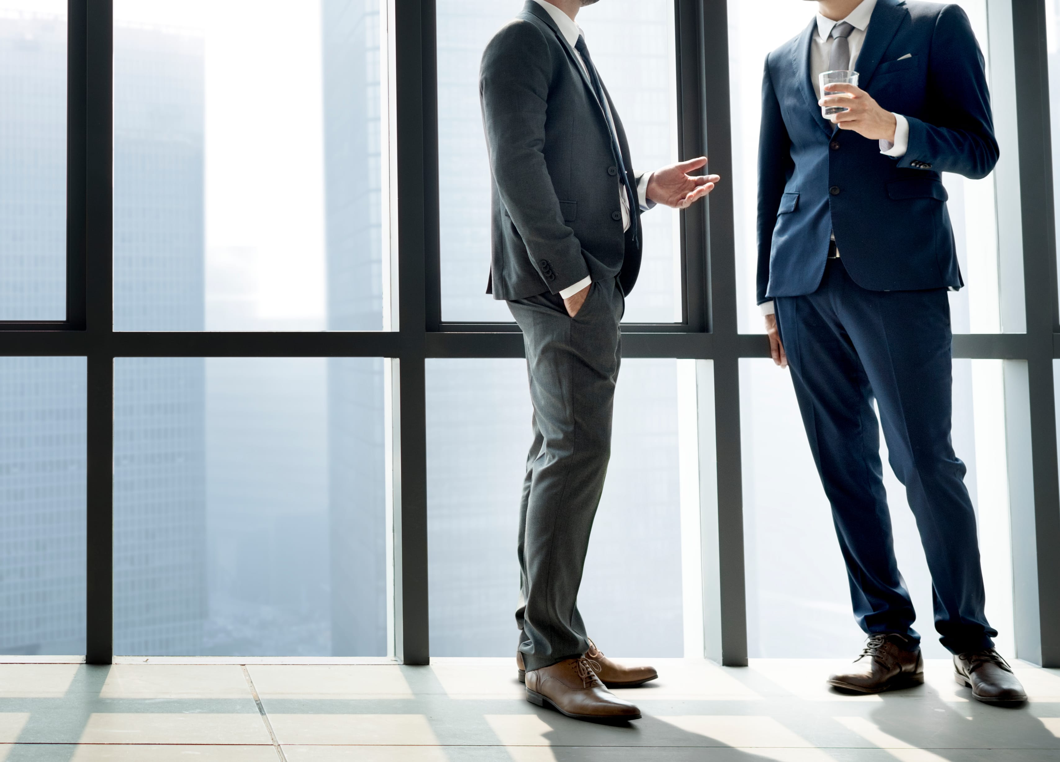 Two men in suits are standing and conversing by large windows; one is holding a glass of water.