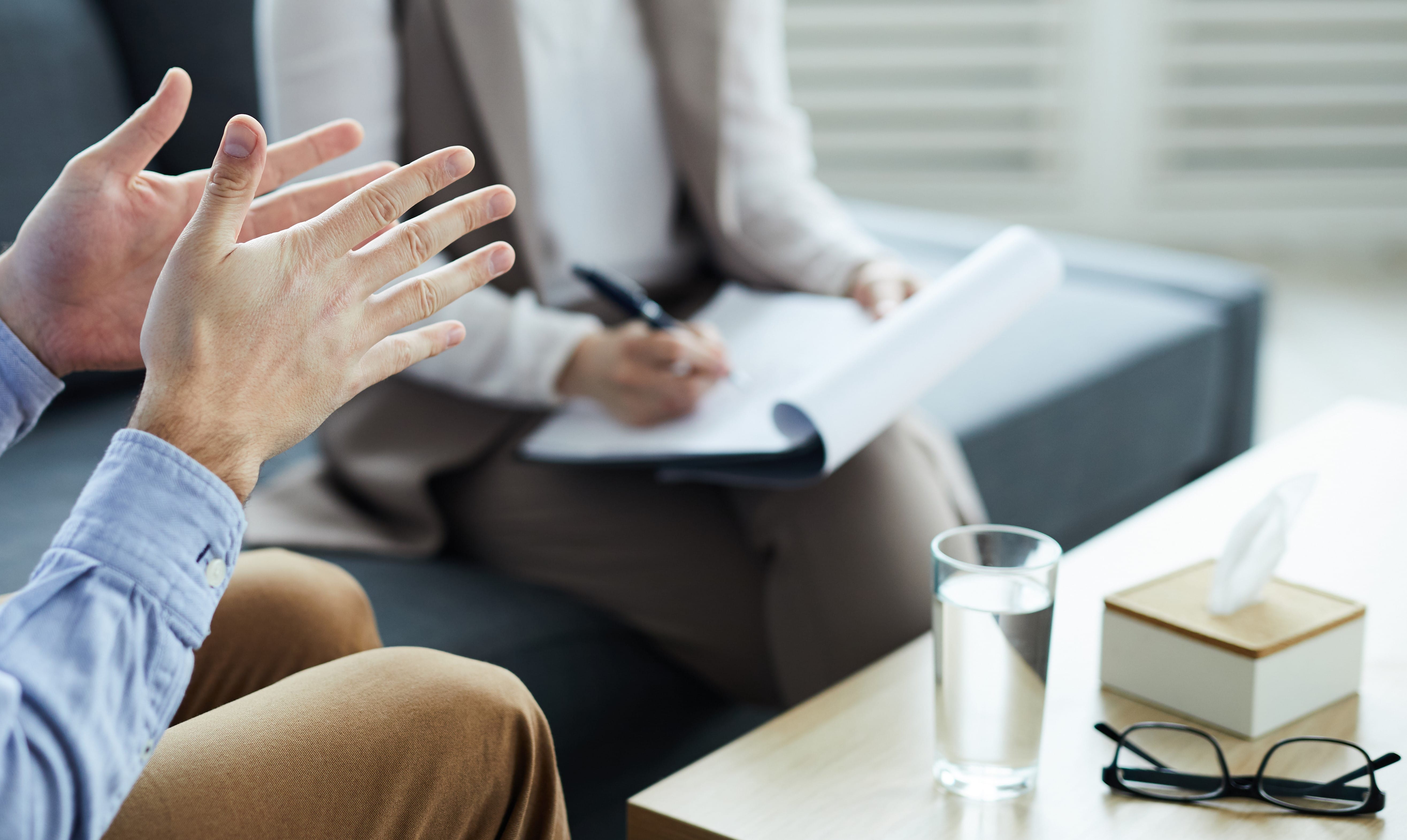 Two people in a discussion, one with gesturing hands, the other writing notes; glasses, tissue box, and water on the table.