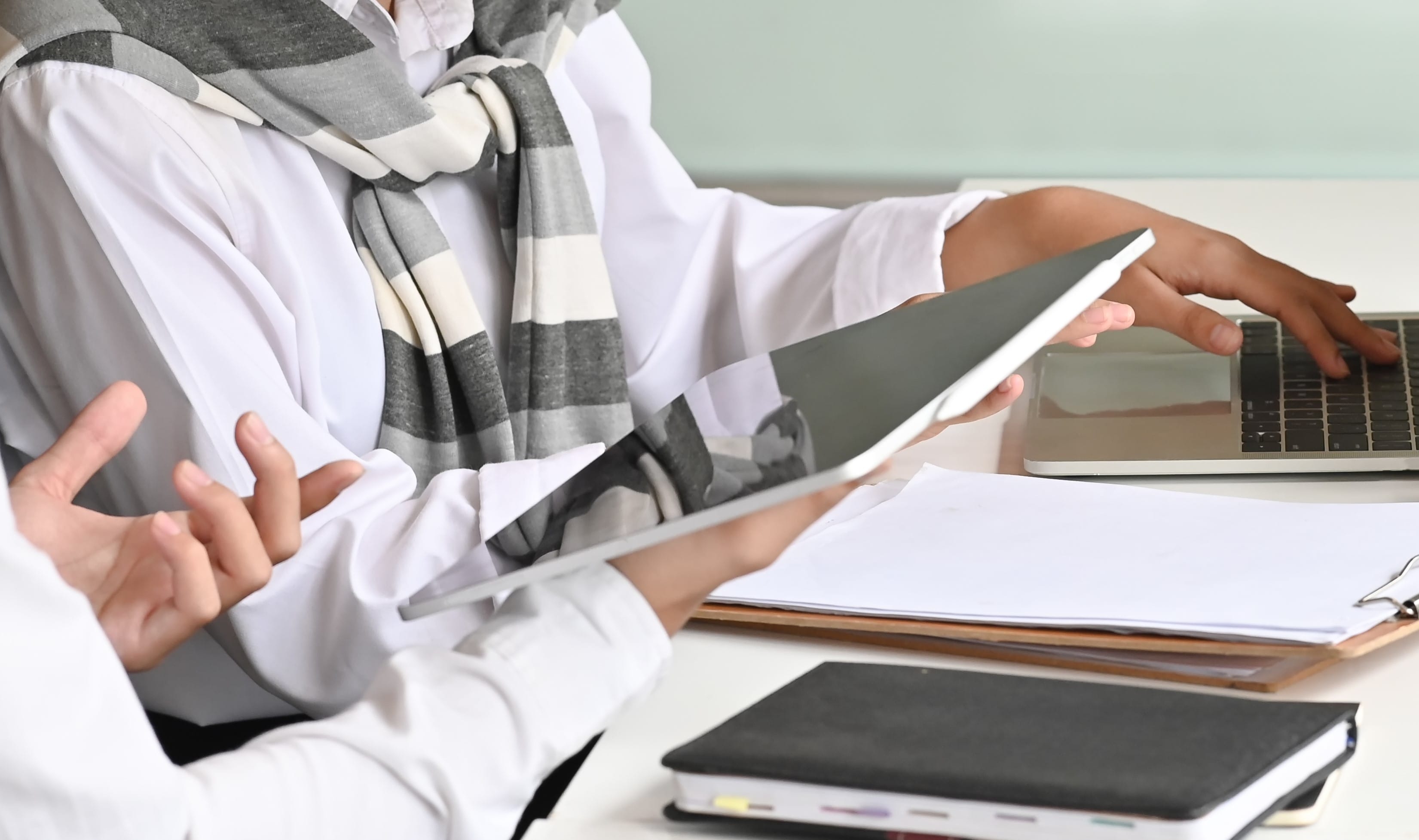 Two people are at a desk, one holding a tablet and the other typing on a laptop, with papers and books nearby.