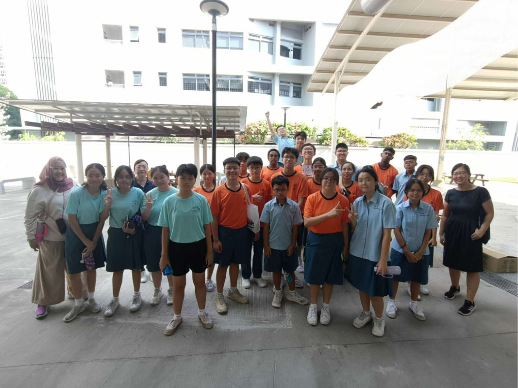 A large group of students in teal and orange uniforms pose outdoors at a school with two adults on either side.