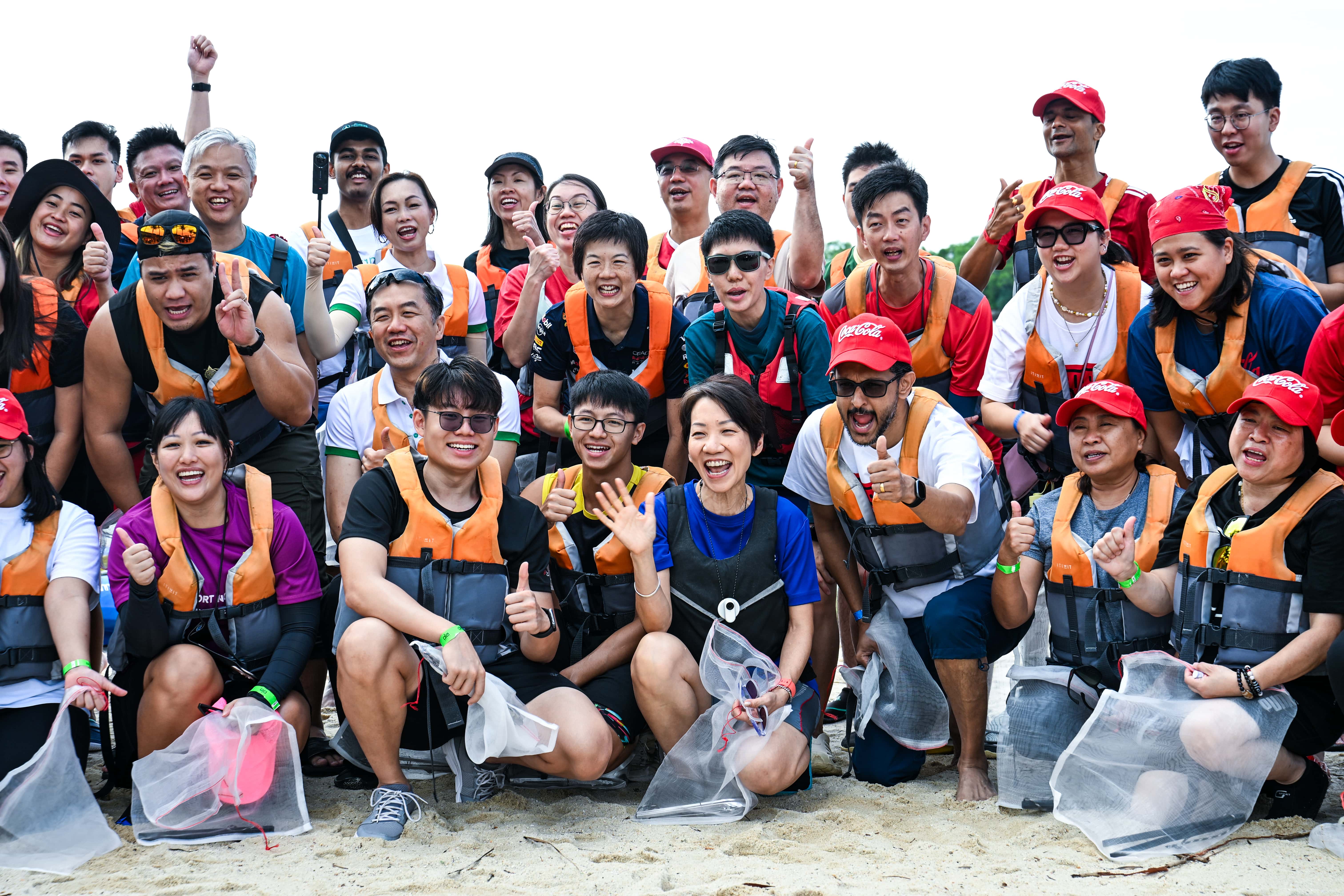 Large group of smiling people wearing life vests pose on a sandy beach, some holding mesh bags.