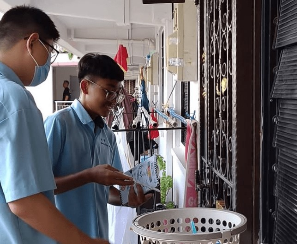 Two boys in blue shirts and masks stand at an apartment door, one holding papers and a basket nearby.