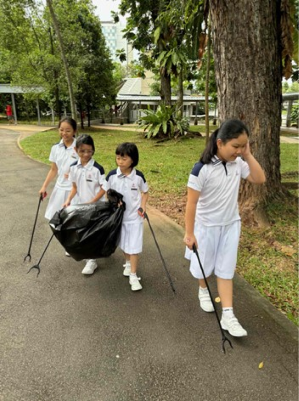 Four children in white uniforms walk on a path holding litter pickers and a black garbage bag.