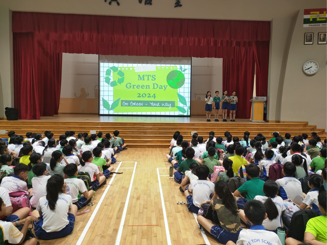 Students sit on a hall floor facing a stage with a screen showing MTS Green Day 2024 Go Green Your Way.