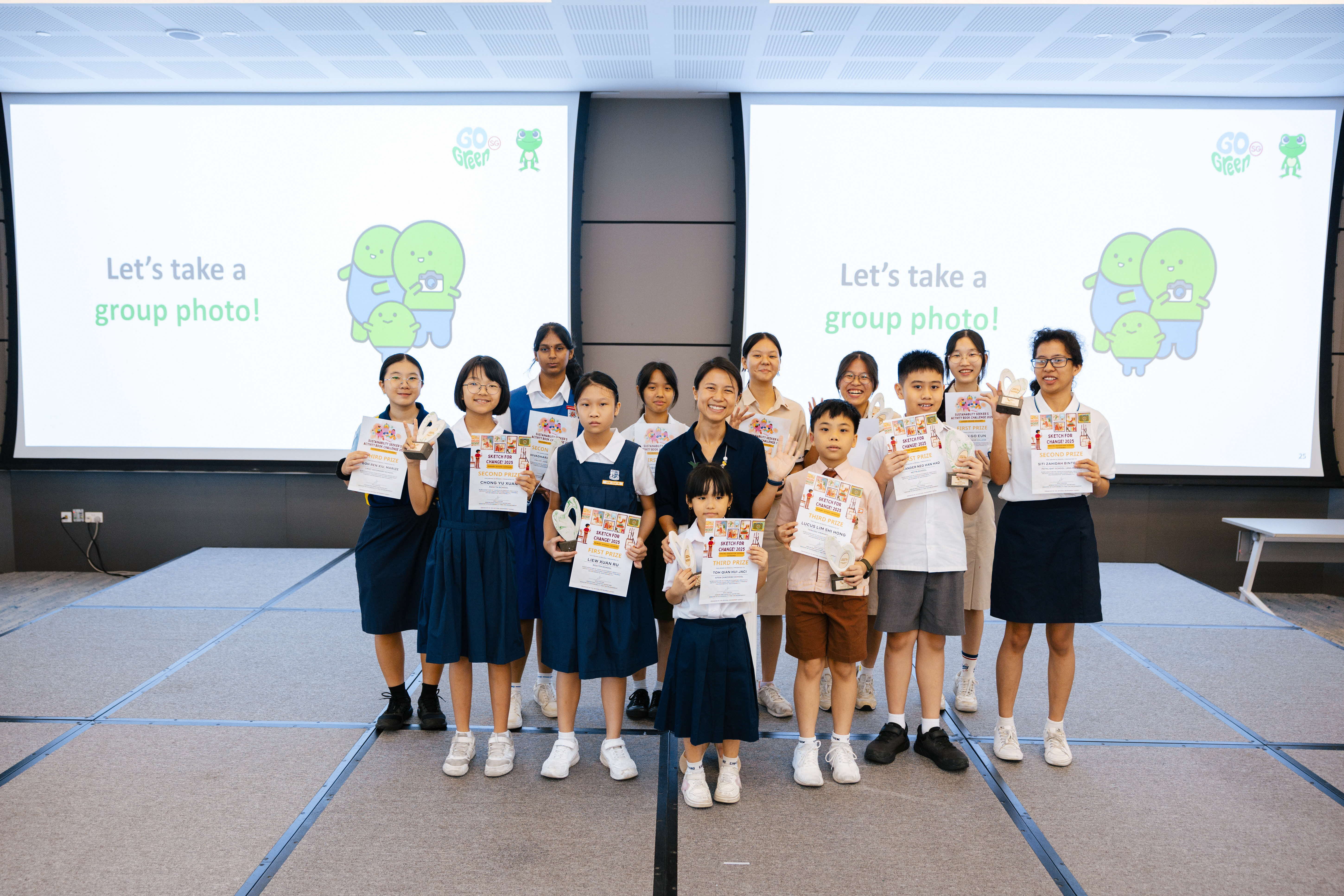 A group of students in school uniforms holding certificates and trophies pose in front of Go Green event screen.