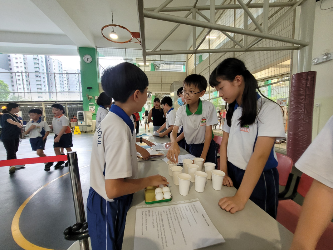 Students in school uniforms stand around a table with paper cups, eggs, and a worksheet in a gymnasium.