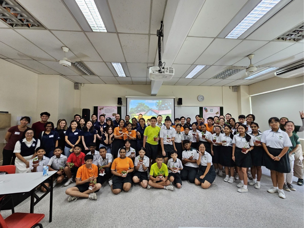 A large group of students and adults pose together in a classroom, many holding small potted plants.