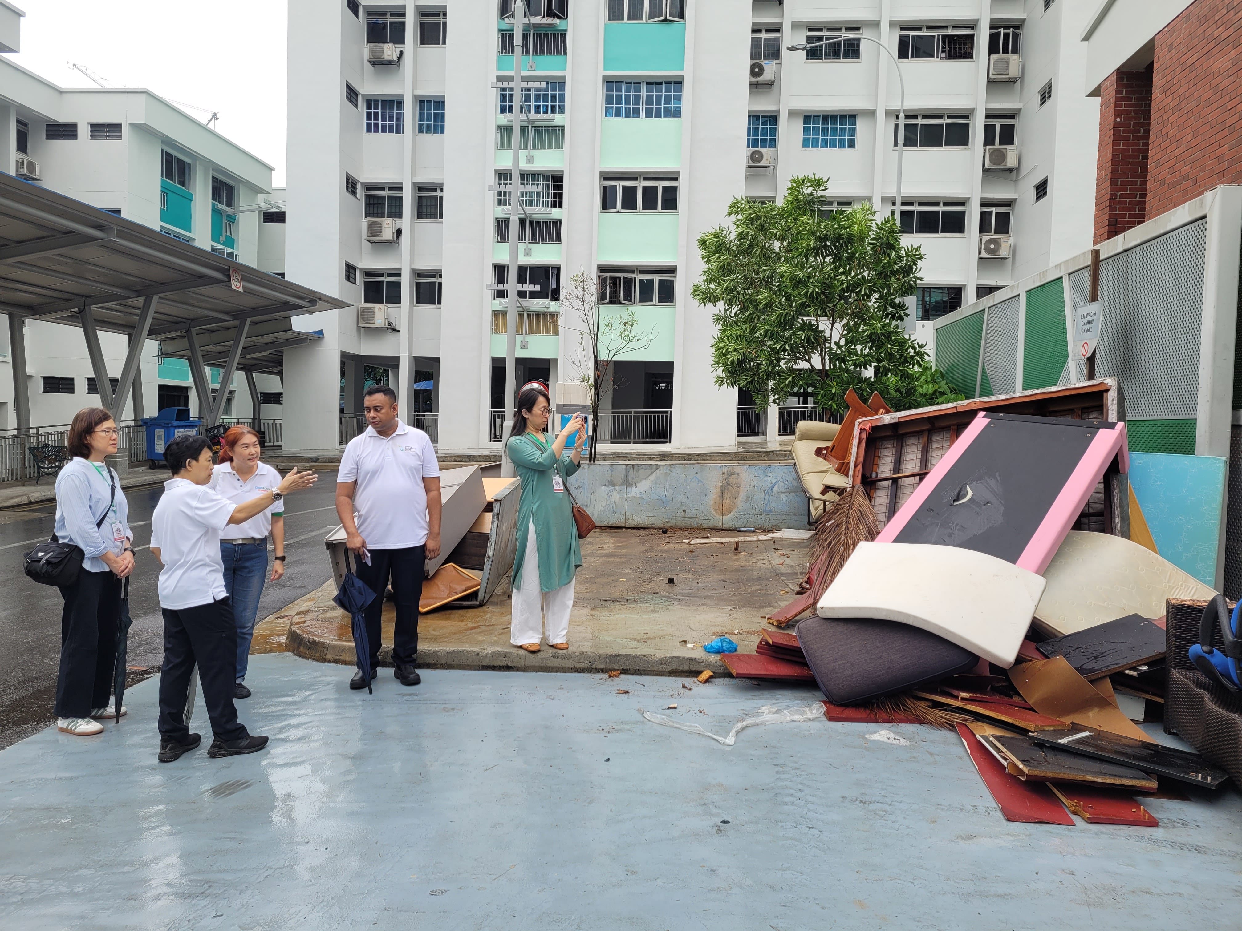 Five people stand near a large pile of discarded furniture and debris outside a tall apartment building.