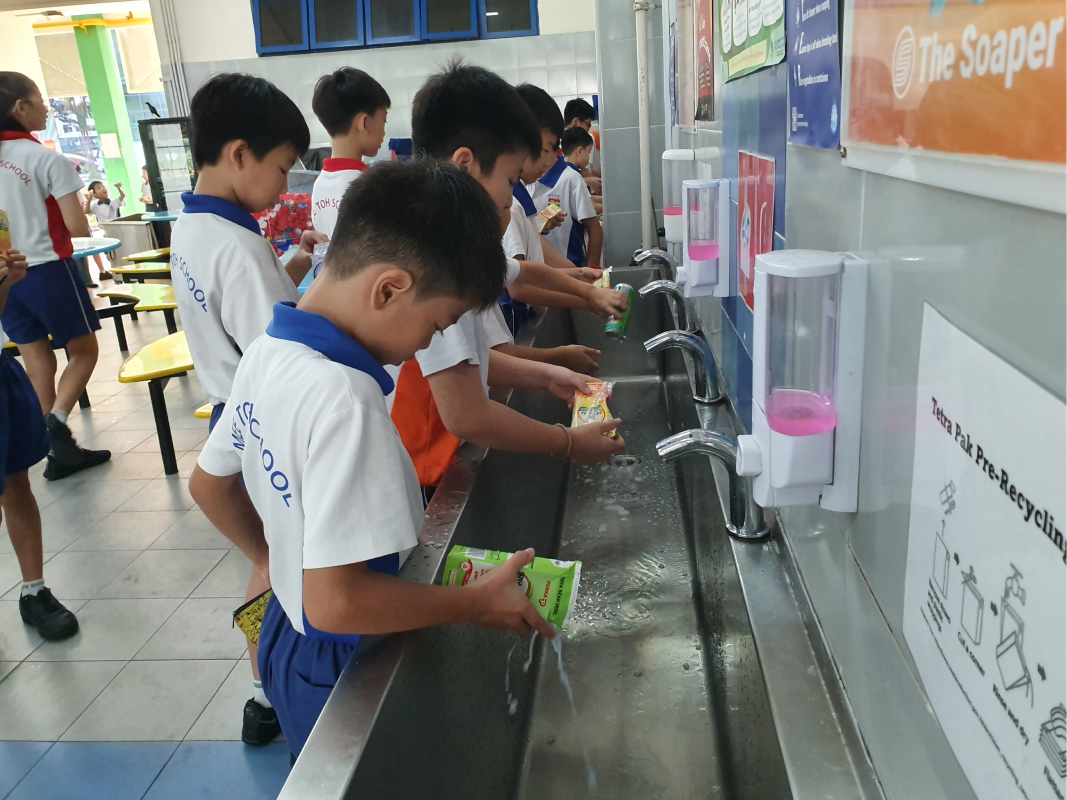 Several school children in uniforms rinse juice cartons at a row of sinks near a Tetra Pak pre-recycling station.
