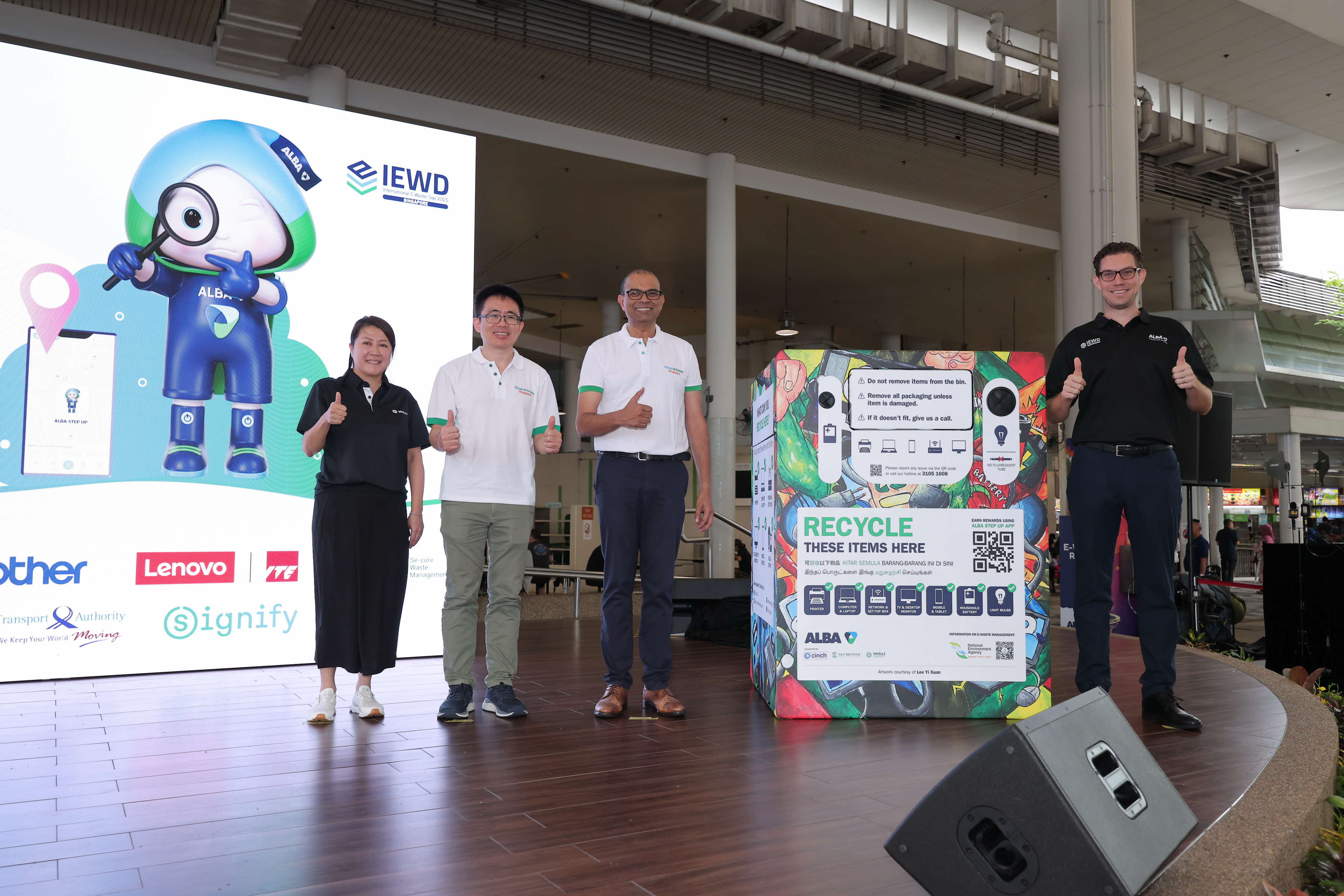 Guests and Minister give thumbs up beside a colorful ALBA e-waste recycling bin at an indoor event with a branded screen.