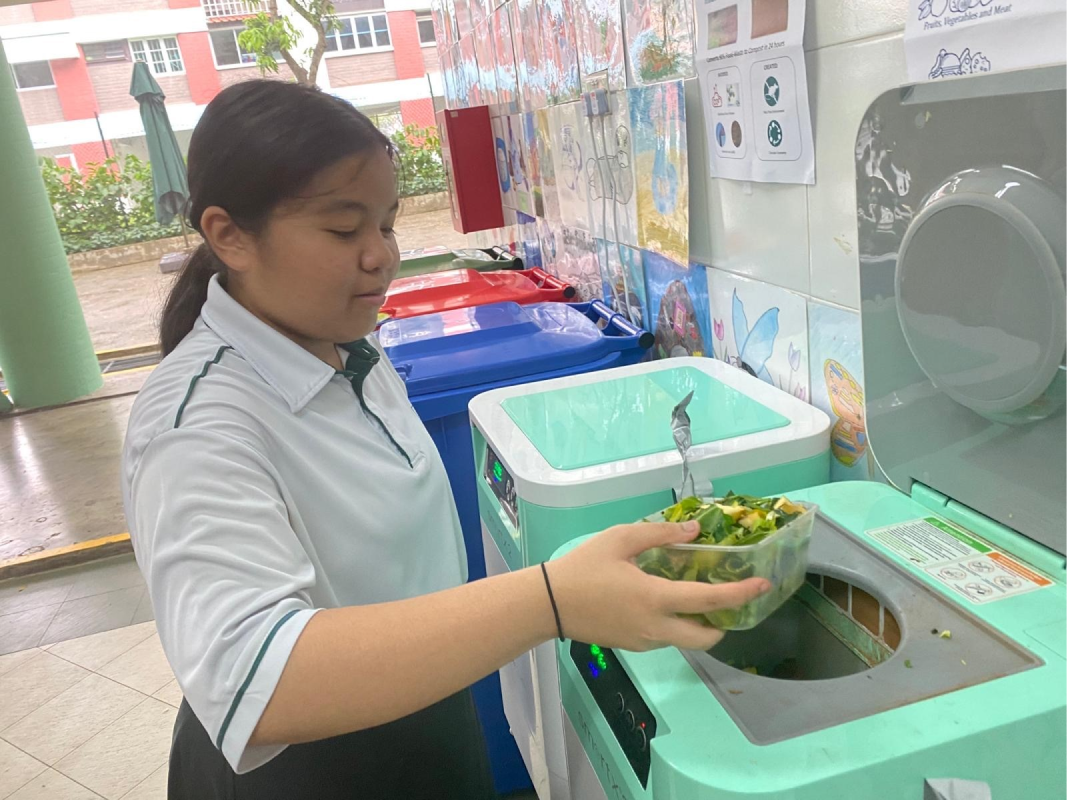 A young girl in school uniform deposits food scraps into a green composting machine near colored recycling bins.