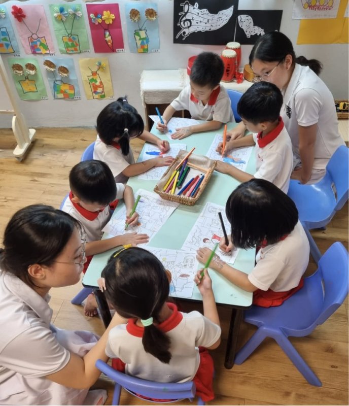 Several young children in uniforms color drawings at a table with two female teachers guiding them in a decorated classroom.
