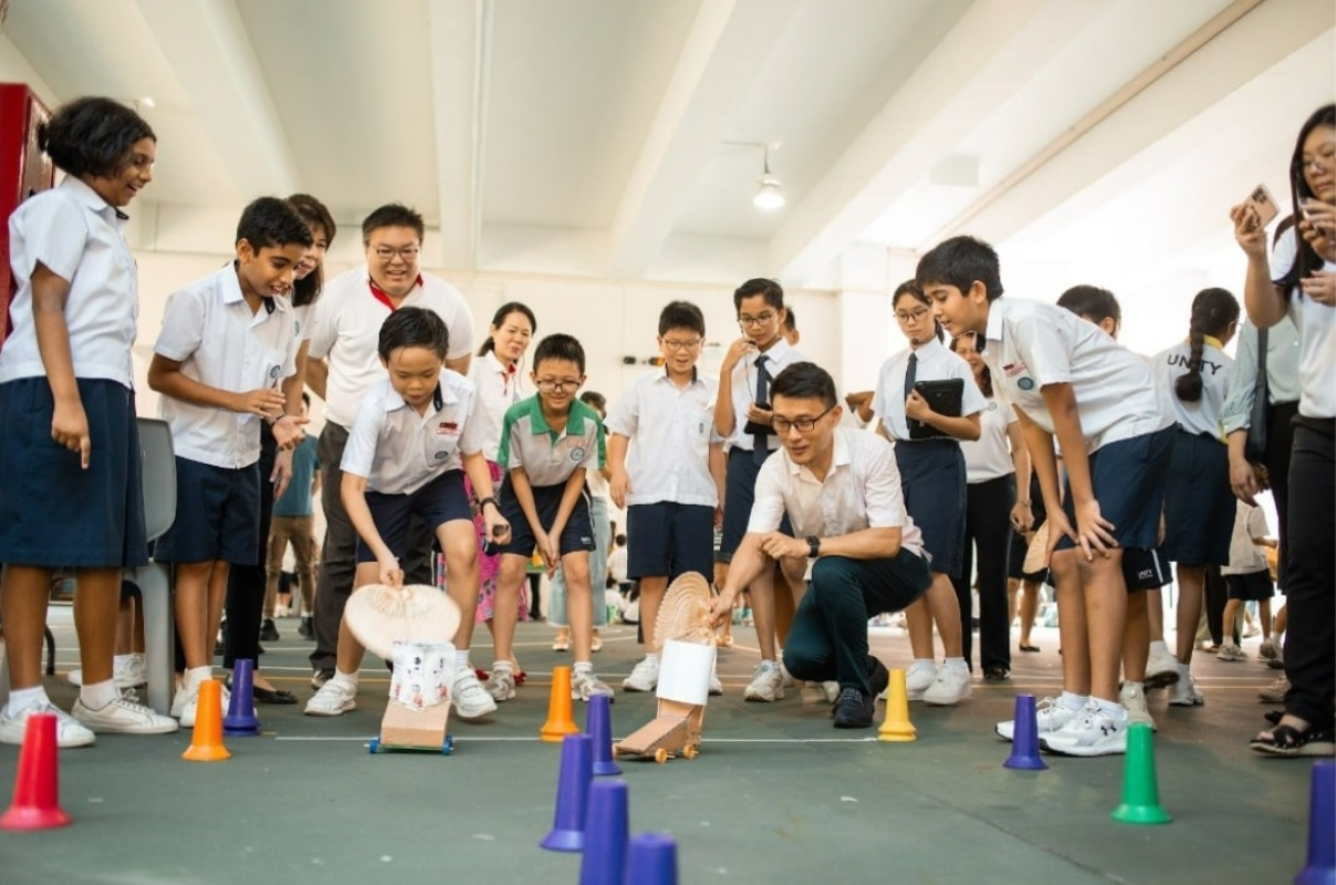 Students in white uniforms race handmade vehicles through colorful cones on a hall floor, watched by a crouching adult.