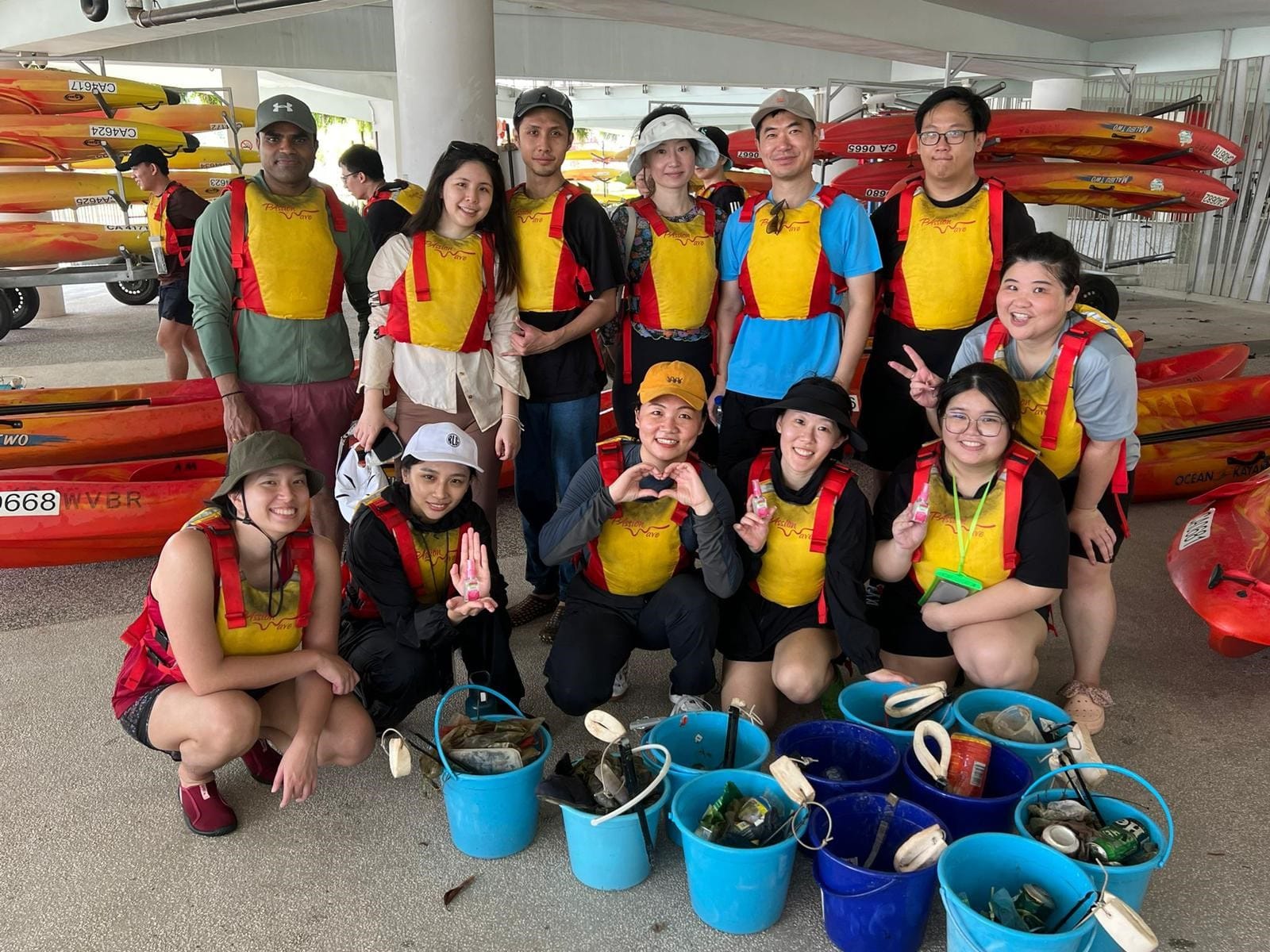 Group of people in life vests posing with buckets of collected trash near stacked red kayaks.