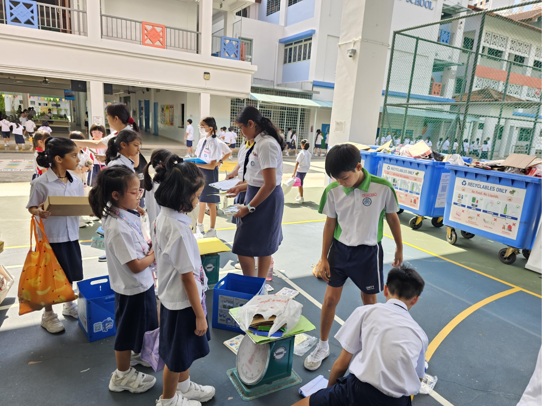Students in uniforms participate in a school recycling drive, weighing items near blue recycling bins outdoors.