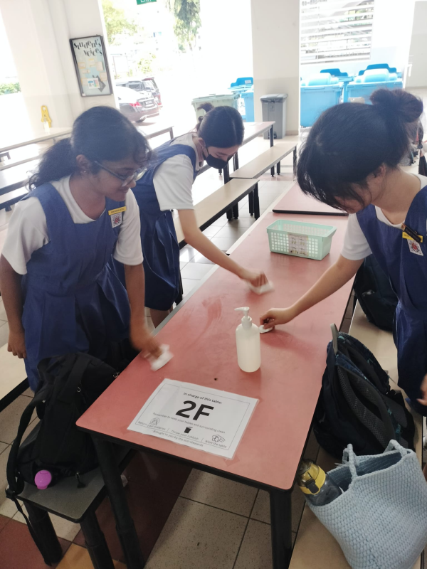 Three schoolgirls in blue uniforms wipe down a table labeled 2F with a sanitizer bottle nearby.