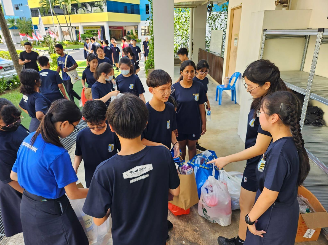 Students in navy blue uniforms gather outdoors near bags of donated goods beside a colorful school building.