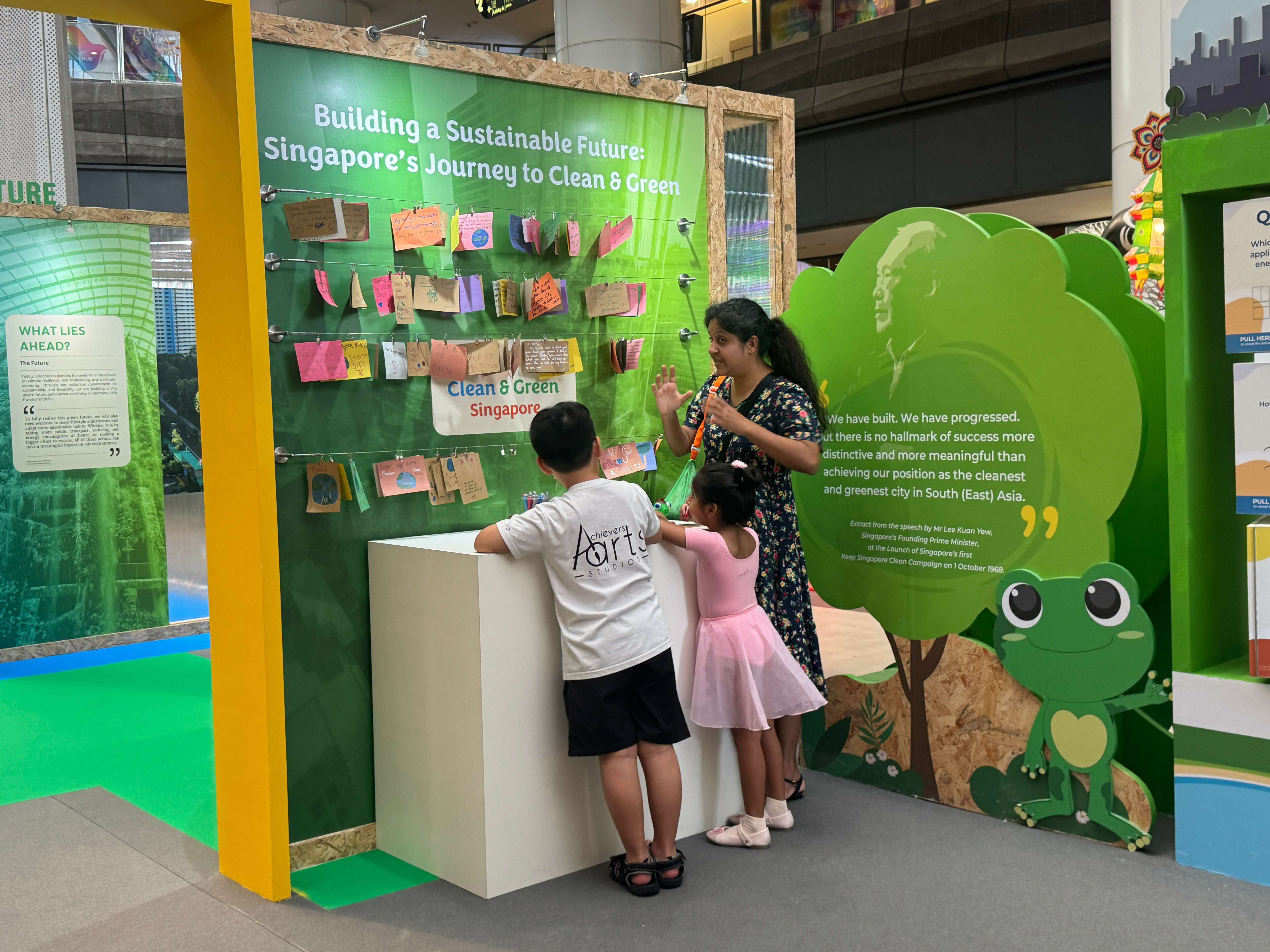 A woman and two children stand at a green exhibit titled "Building a Sustainable Future: Singapore's Journey to Clean & Green."