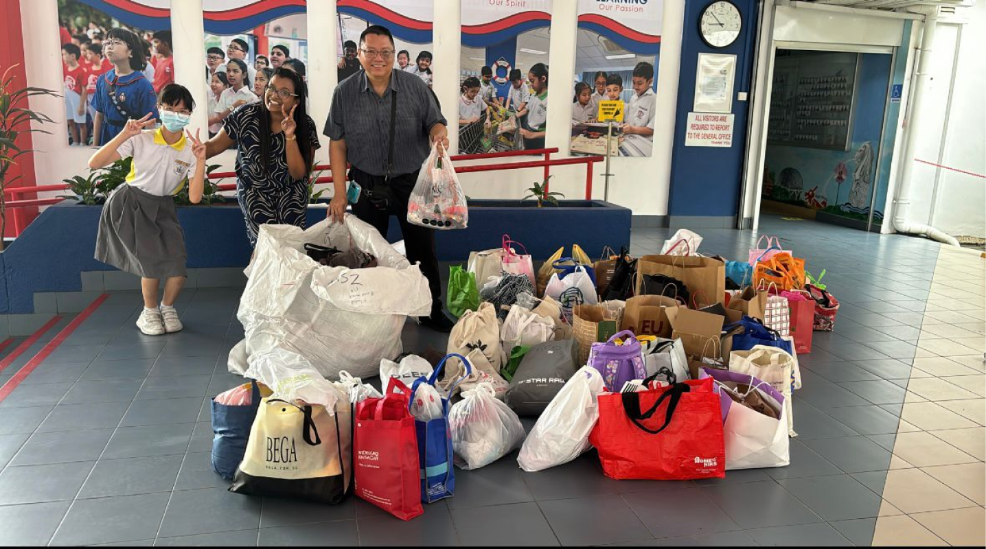 Two adults stand smiling behind a large collection of donation bags in a school lobby, with a child nearby.