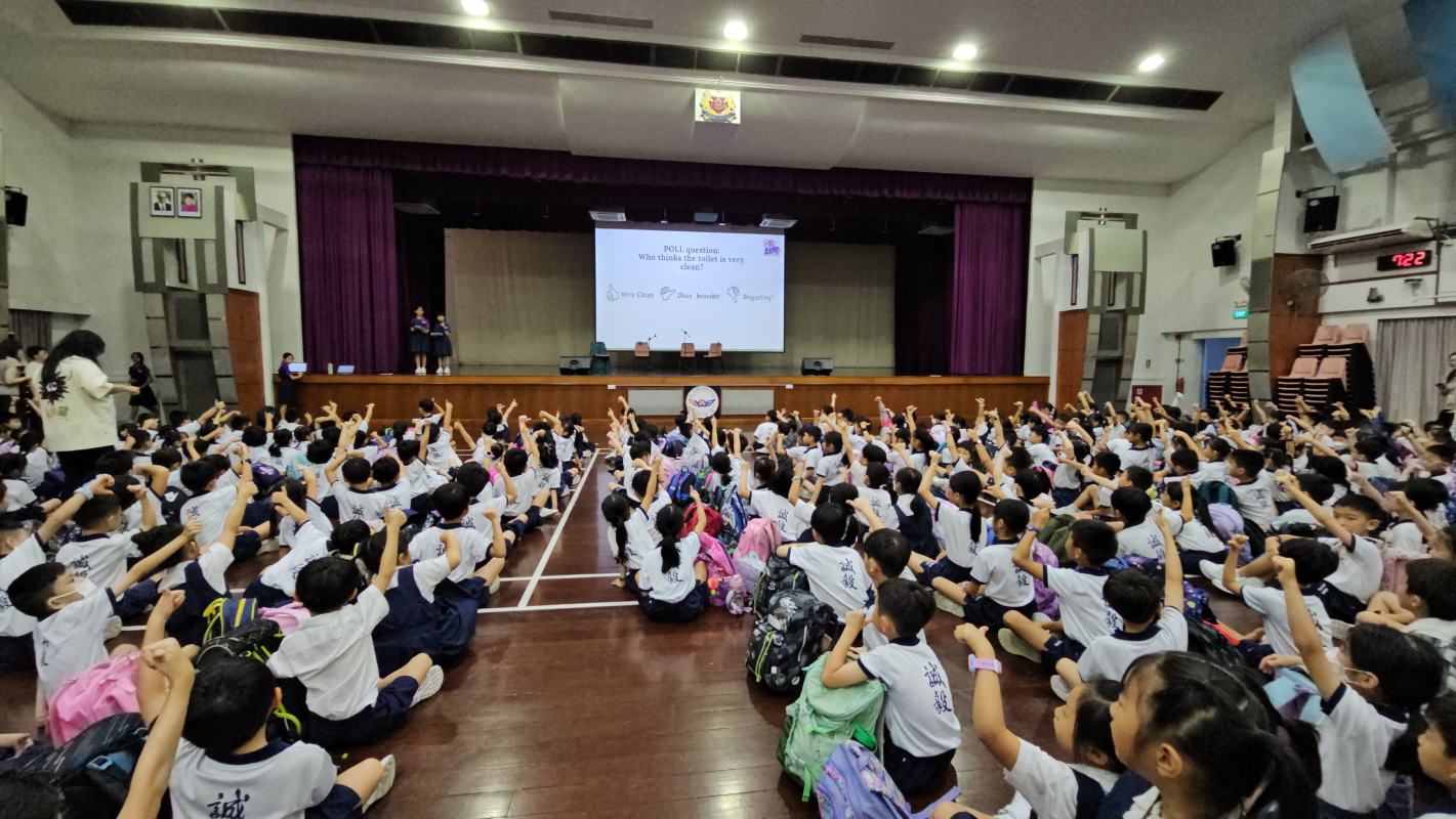 Hundreds of school children in white uniforms sit on a hall floor raising their hands toward a stage with a projected poll slide.