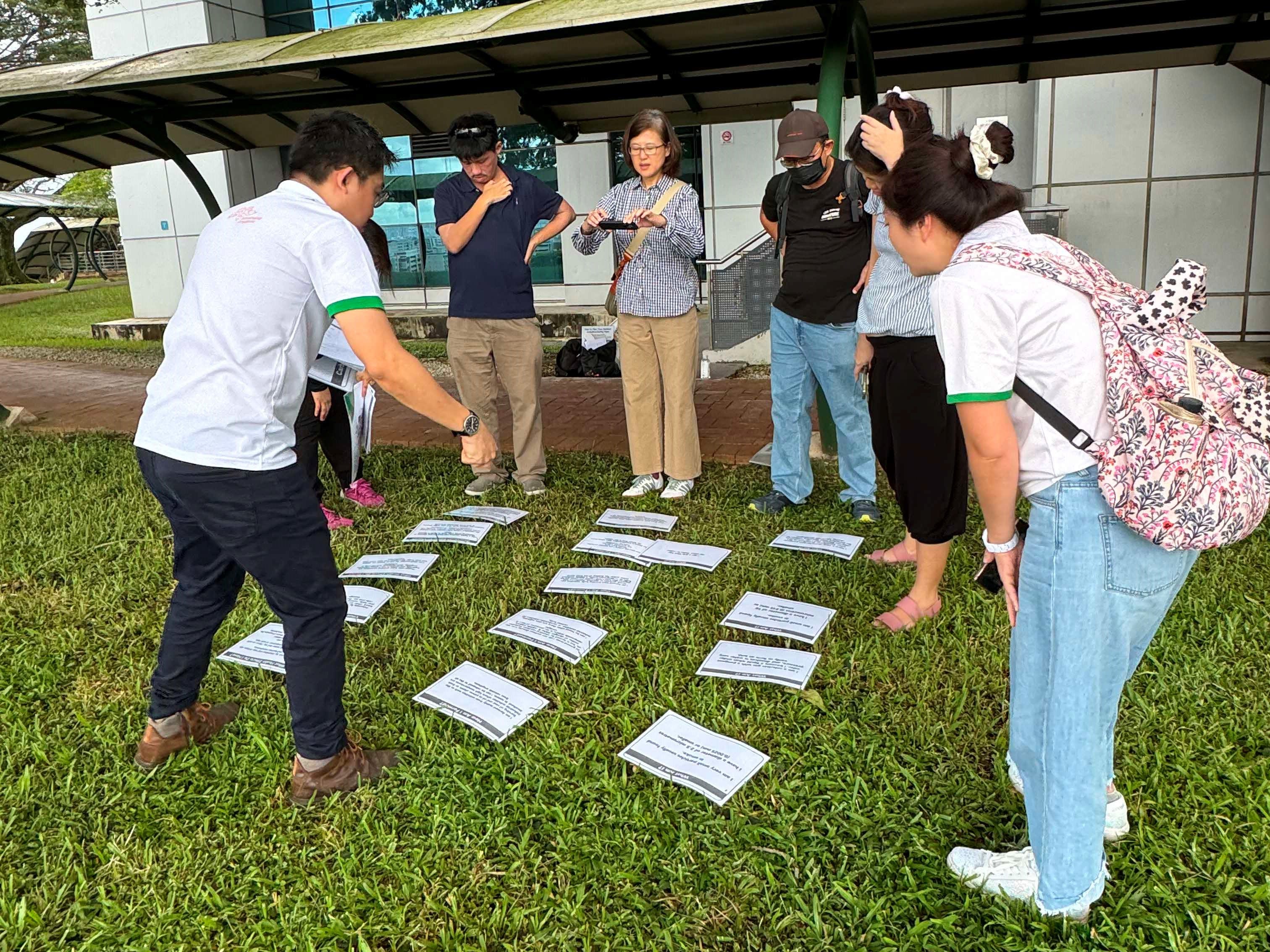 Six people stand on grass outdoors, looking down at printed cards.