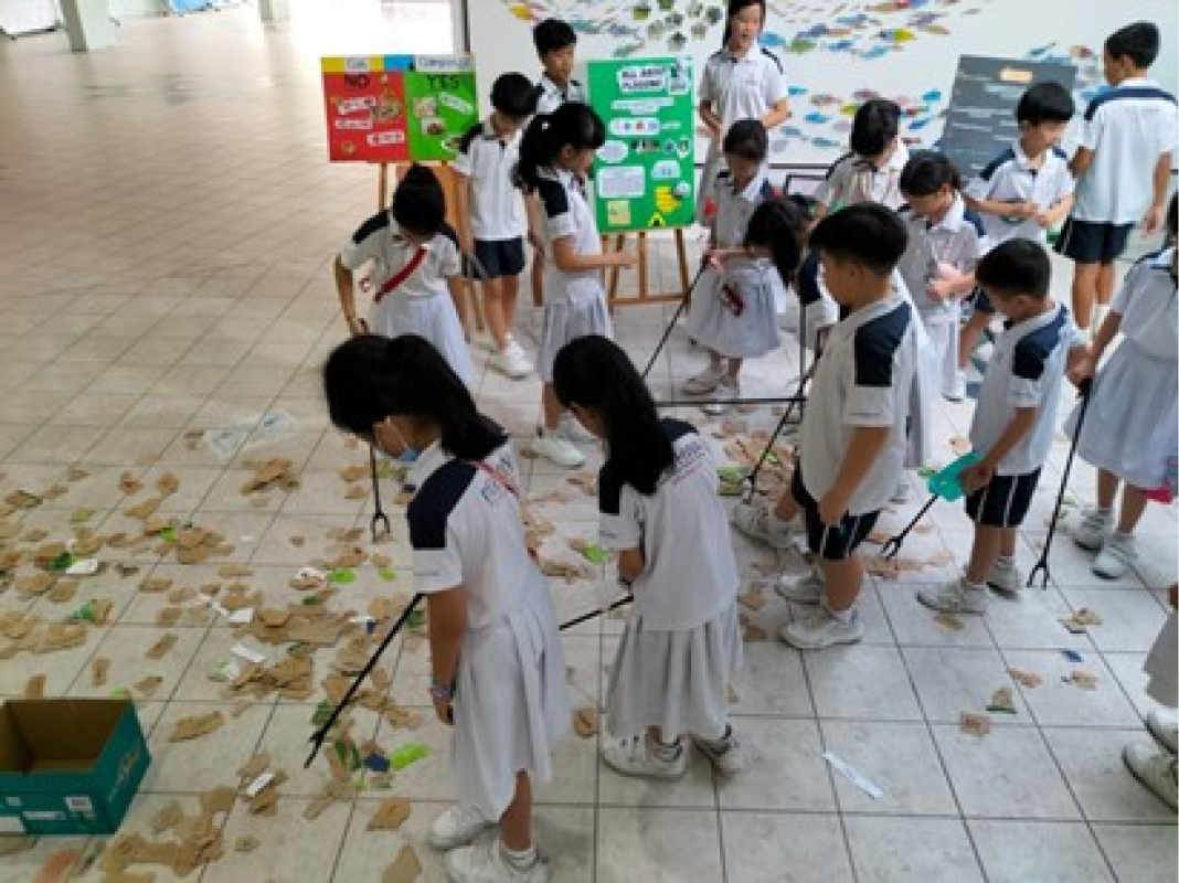 Uniformed schoolchildren use litter pickers to collect debris from a tiled floor near recycling display boards.