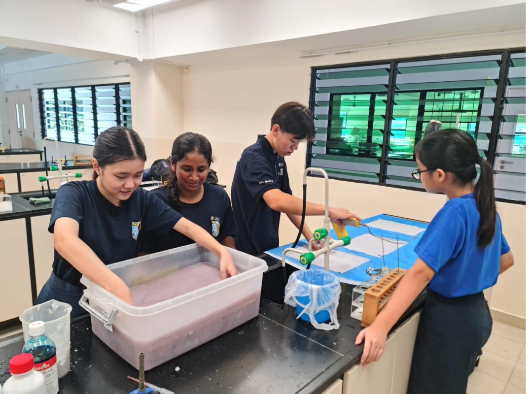 Four students in a school laboratory work around a black bench with a large tray of liquid and lab equipment.