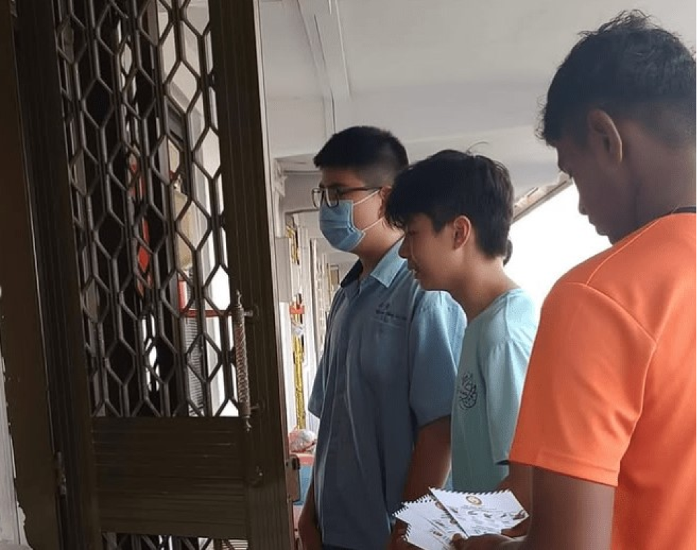 Three young men stand at a metal gate door, one wearing a face mask and glasses, another holding pamphlets.